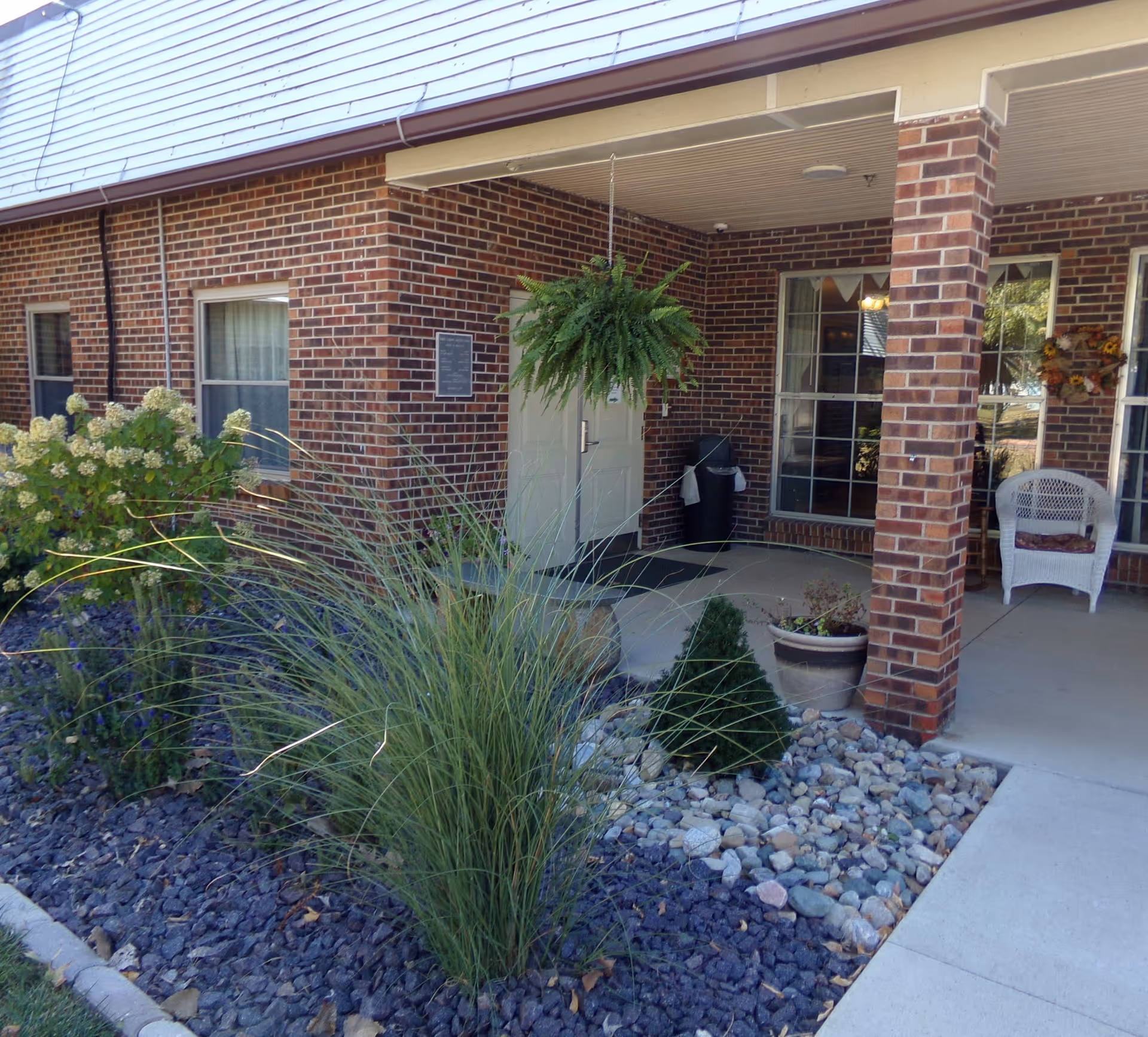 Brick senior living facility entrance with hanging fern, porch chair, and rock landscaping.