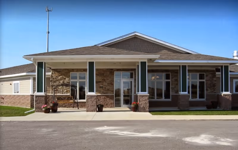 Front exterior view of a single-story building with a covered entrance supported by columns. The building features stone and brick accents, large windows, and a swing bench on the porch. There are potted plants near the entrance and a clear blue sky above.