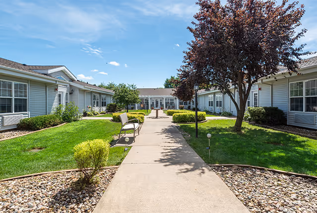 Outdoor courtyard area of Homestead Assisted Living of Denison with a concrete walkway flanked by green grass, bushes, and trees. Benches are placed along the walkway, and single-story buildings with white siding and multiple windows surround the courtyard under a blue sky with some clouds.