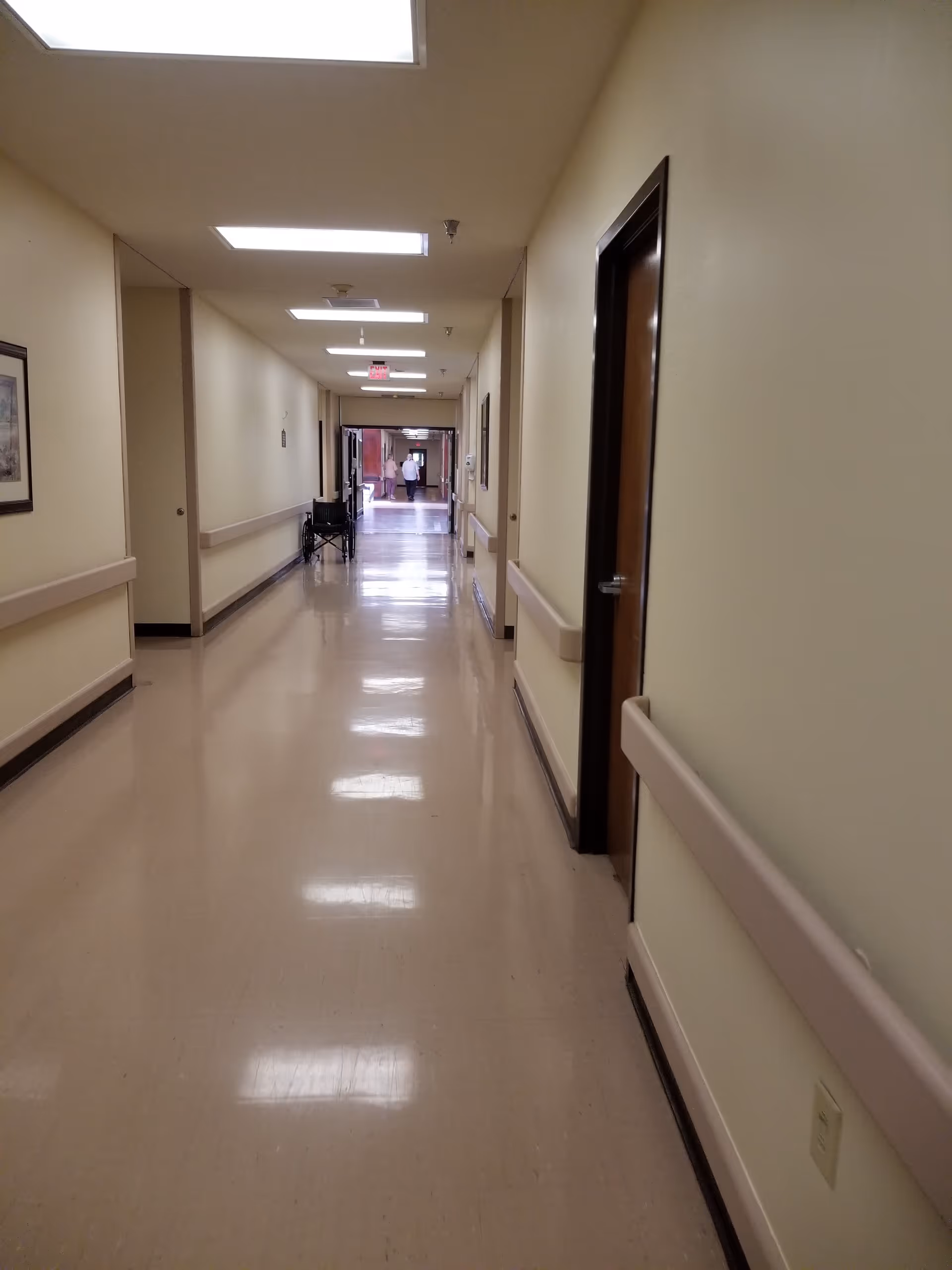 Long beige-walled hallway in a senior living facility with overhead lights and handrails.
