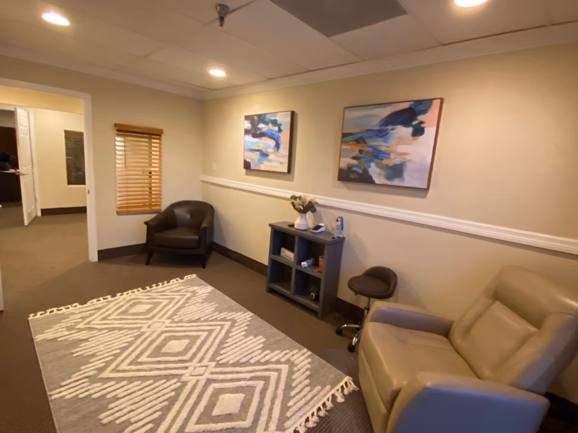 A cozy senior living lounge with tan chairs, wall art, and a geometric rug.