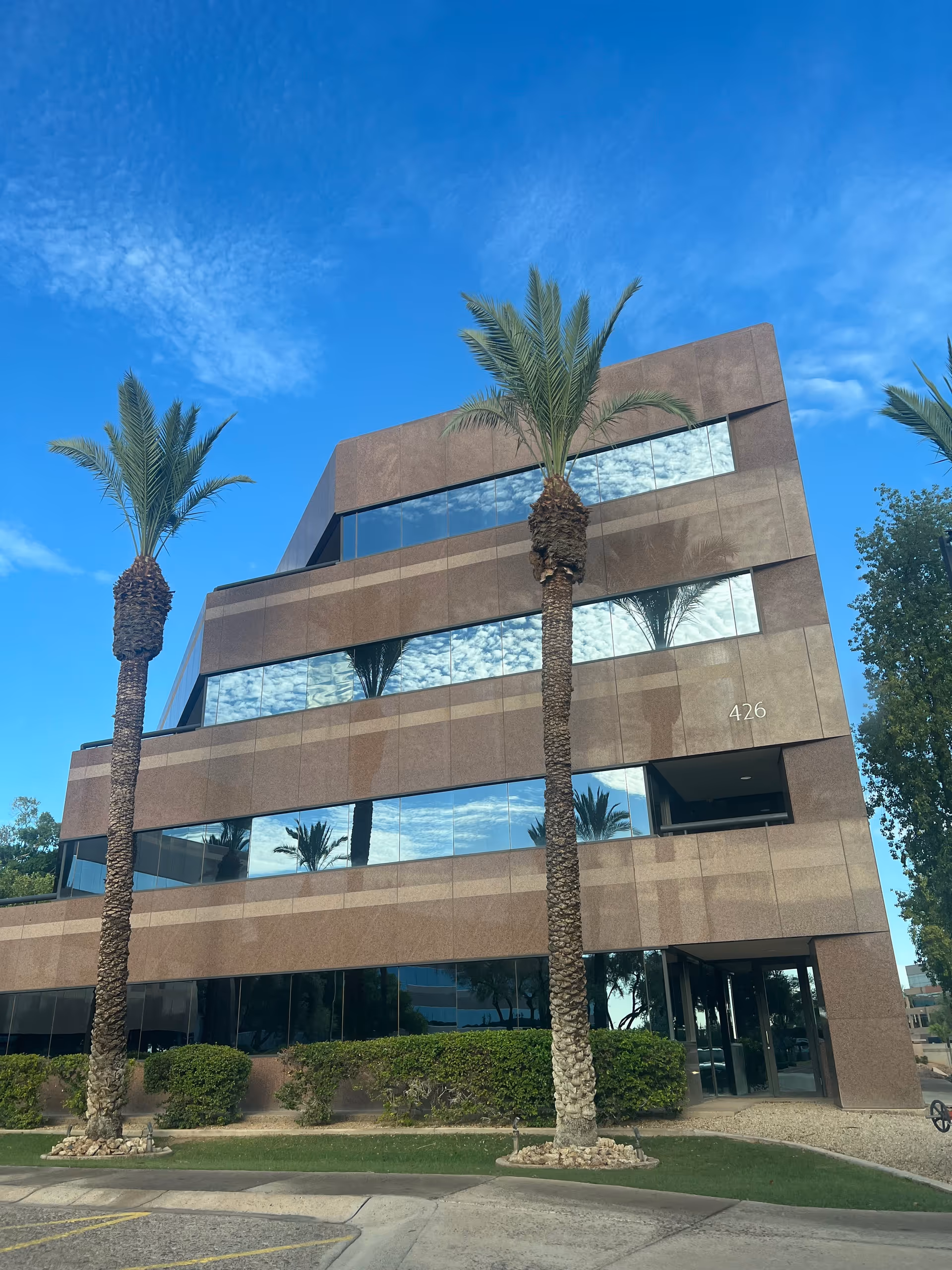 Modern multi-story brown glass building with palm trees and a clear blue sky.