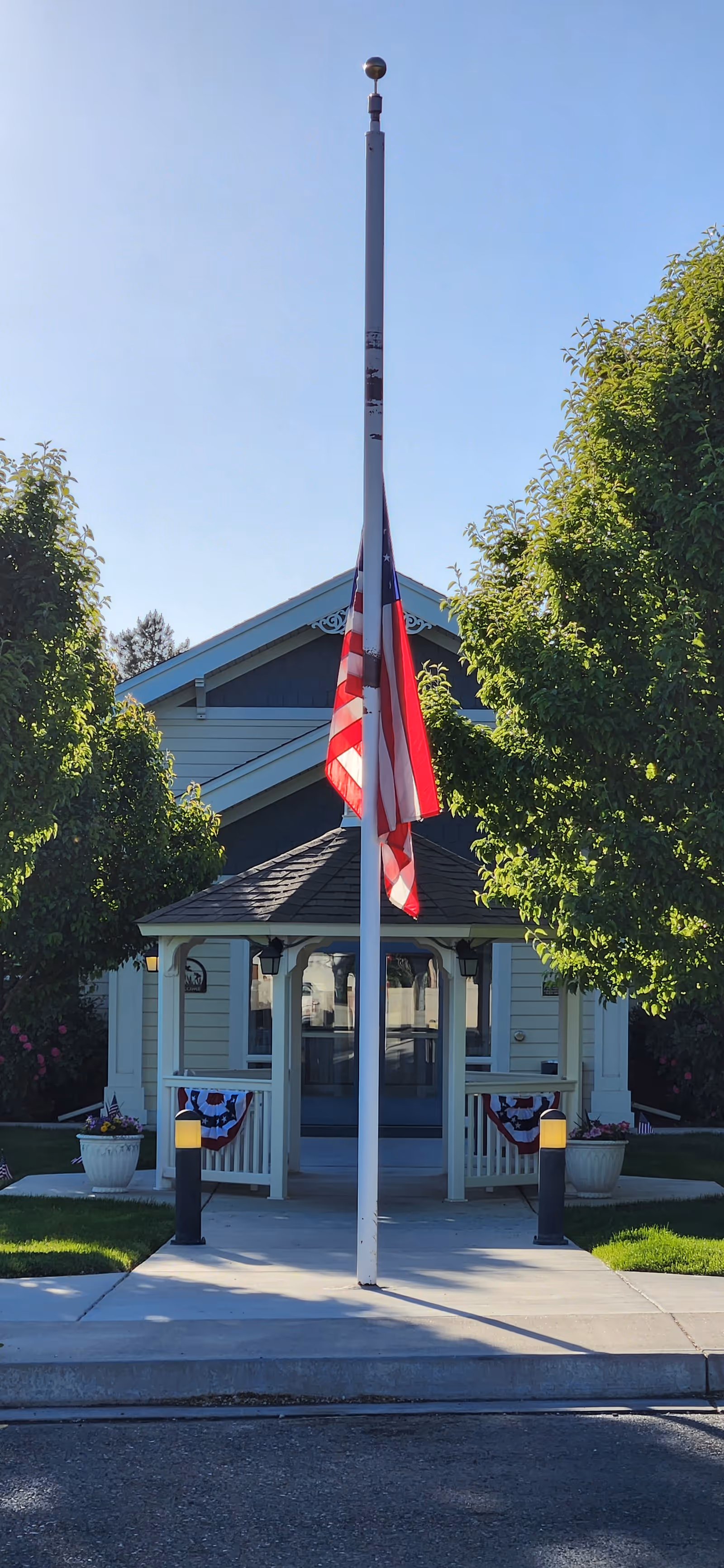 A flagpole with an American flag at half-mast stands in front of a small building with a covered porch entrance. The building is light-colored with a dark roof and is flanked by two green trees. There are decorative planters and patriotic bunting on the porch railing.