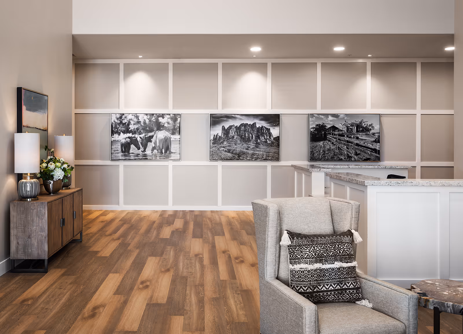 A modern interior common area with wood flooring, a gray armchair with a patterned cushion, a wooden sideboard with two lamps and a flower arrangement, and a white reception desk with granite countertops. The back wall features decorative paneling and three black and white framed photographs depicting horses, a mountain landscape, and a rustic fence.
