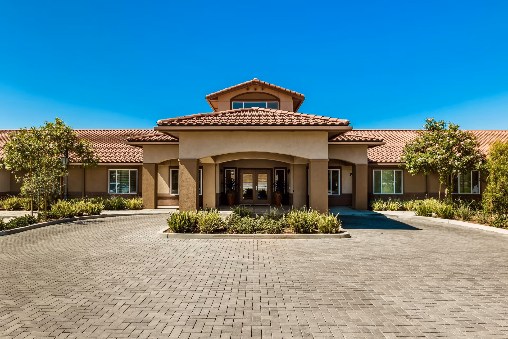 Front exterior view of a single-story building with a tiled roof and a covered entrance. The building is surrounded by neatly trimmed bushes and small trees, with a paved driveway leading up to the entrance under a clear blue sky.