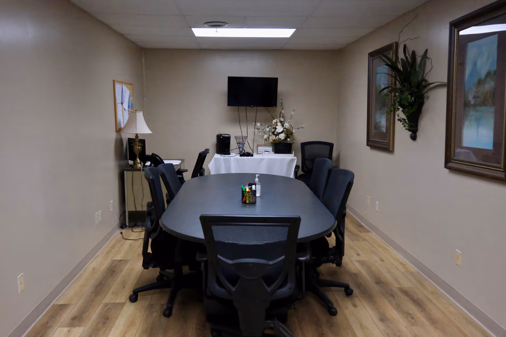 A neutral conference room with an oval table, rolling chairs, and a wall-mounted TV.