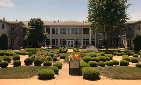 Sunlit courtyard with round manicured shrubs, flower planters, and a two-story assisted living building across a central walkway.