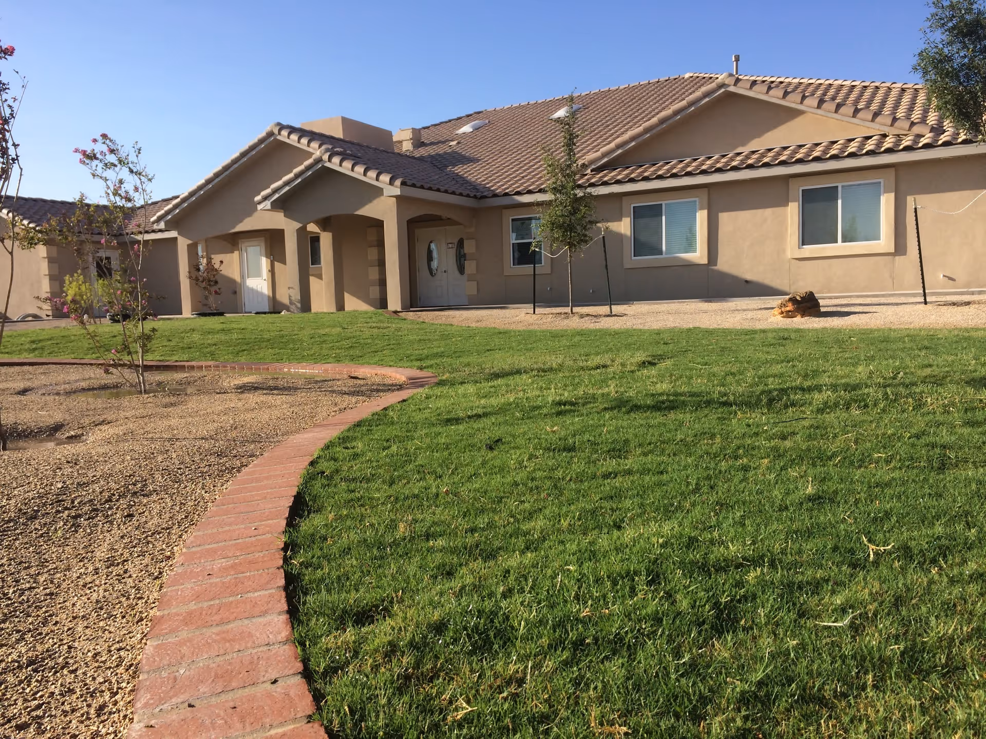 Single-story building with beige stucco walls and a tiled roof, surrounded by a well-maintained lawn and a curved brick border separating the grass from a gravel area with small trees and shrubs under a clear blue sky.