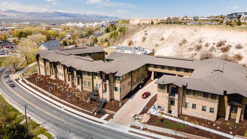 Aerial view of a large senior living facility building with multiple wings and a driveway entrance. The building has a brown roof and beige exterior walls. Surrounding the facility are roads, some greenery, and a hillside in the background with a cityscape and mountains visible in the distance under a partly cloudy sky.