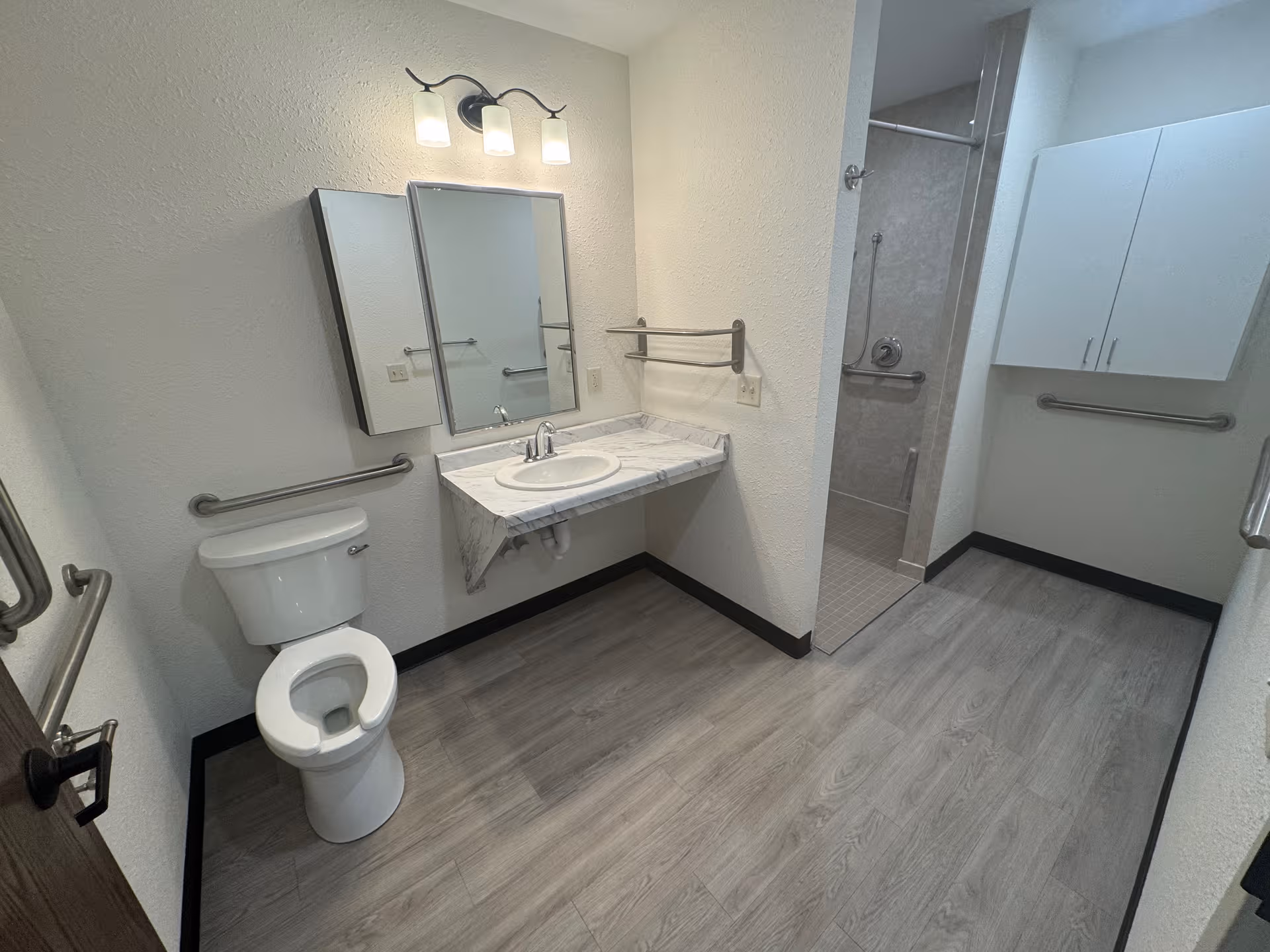 Accessible bathroom with toilet, sink, mirrors, grab bars, and a tiled shower stall.