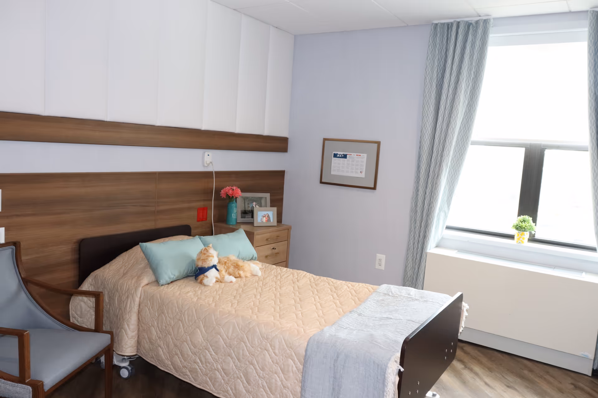 A bedroom in a senior living facility with a bed, nightstand, pillows, and window.