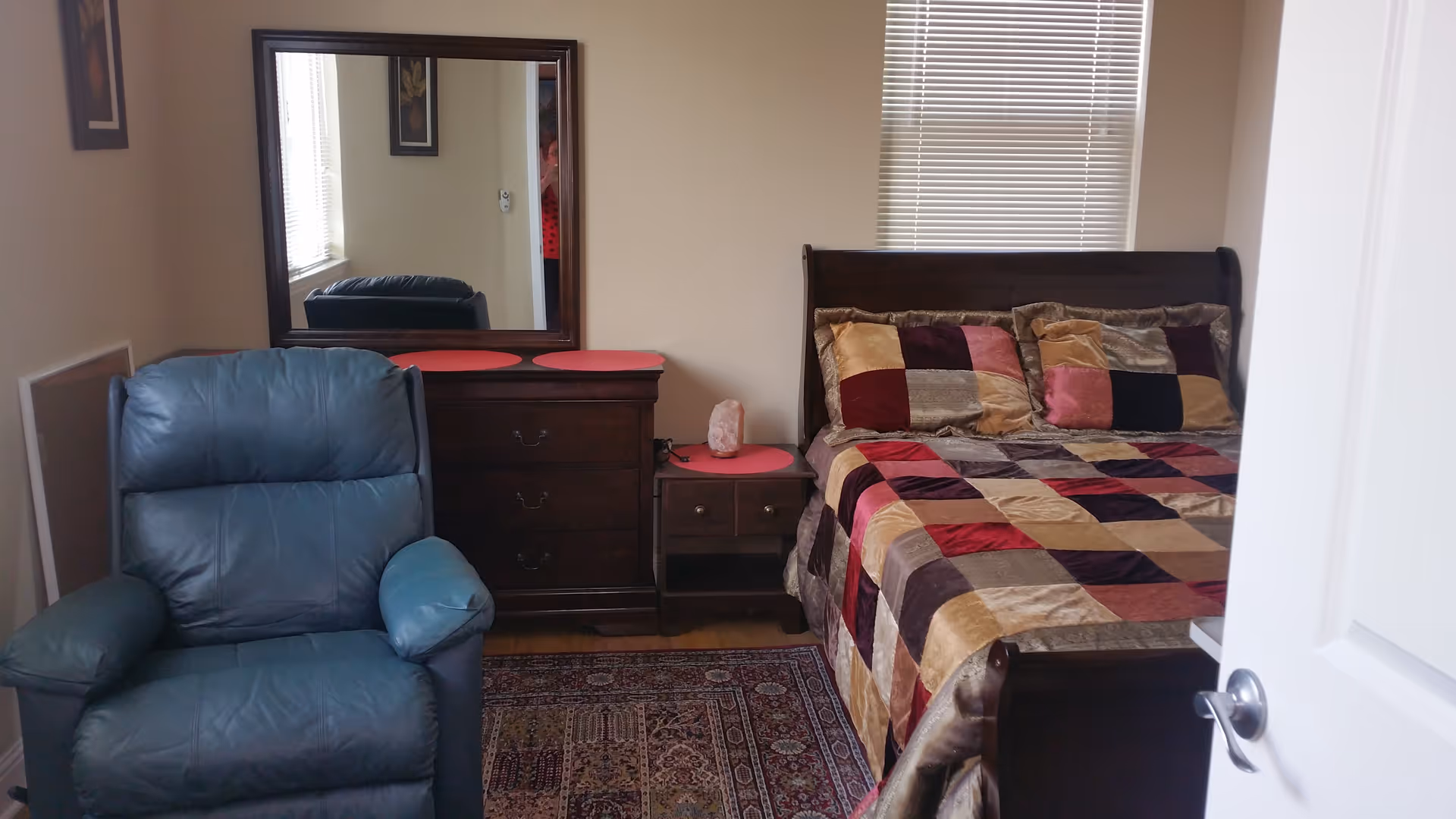 A cozy bedroom with a wooden bed featuring a colorful patchwork quilt and matching pillows. Next to the bed is a wooden nightstand with a lamp, and a wooden dresser with a large mirror above it. A blue leather armchair is positioned near the dresser. The room has beige walls, a window with blinds, and a patterned rug on the floor.