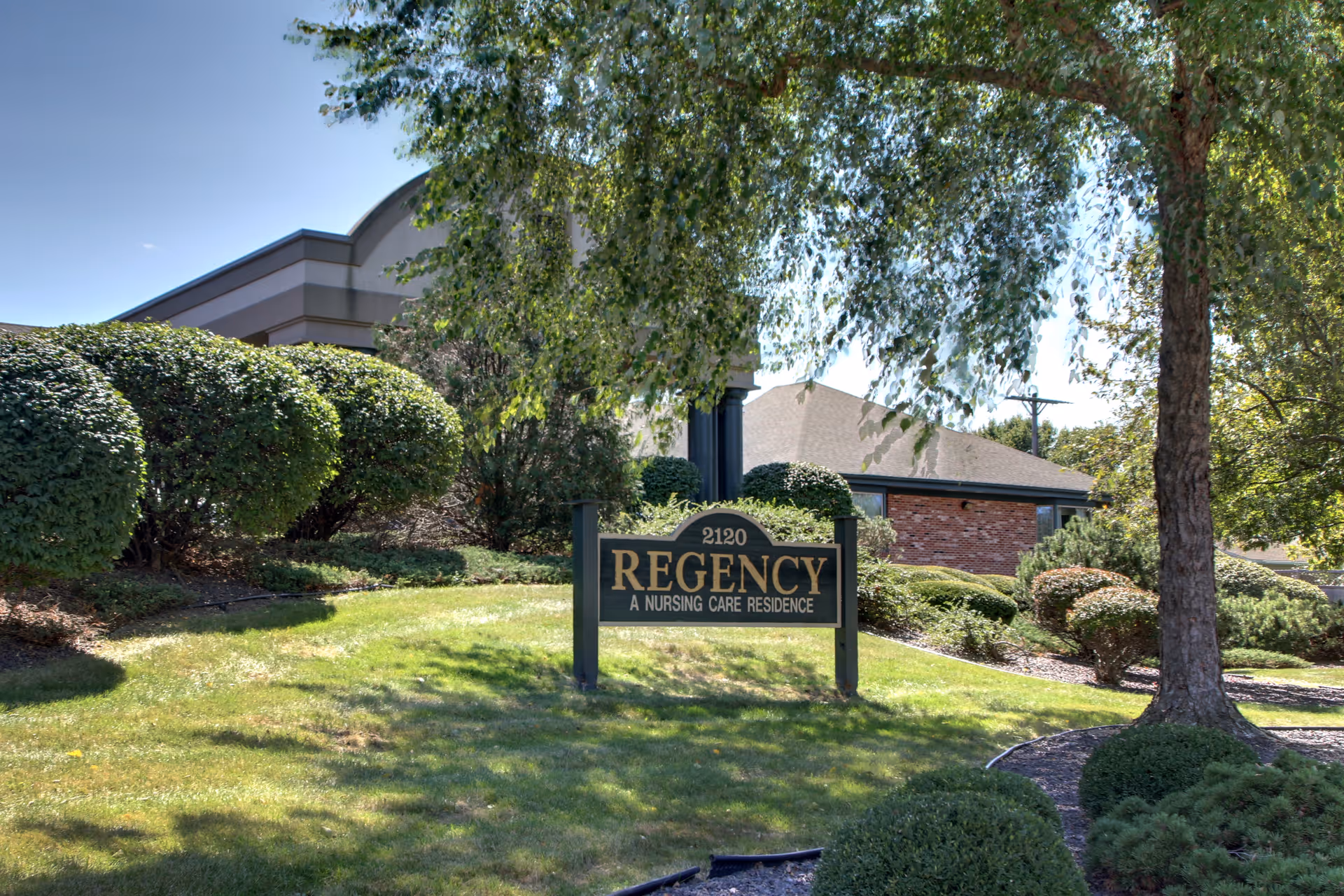 Outdoor view of the Regency nursing care residence sign surrounded by well-maintained bushes, trees, and grass with part of the building visible in the background under a clear blue sky.