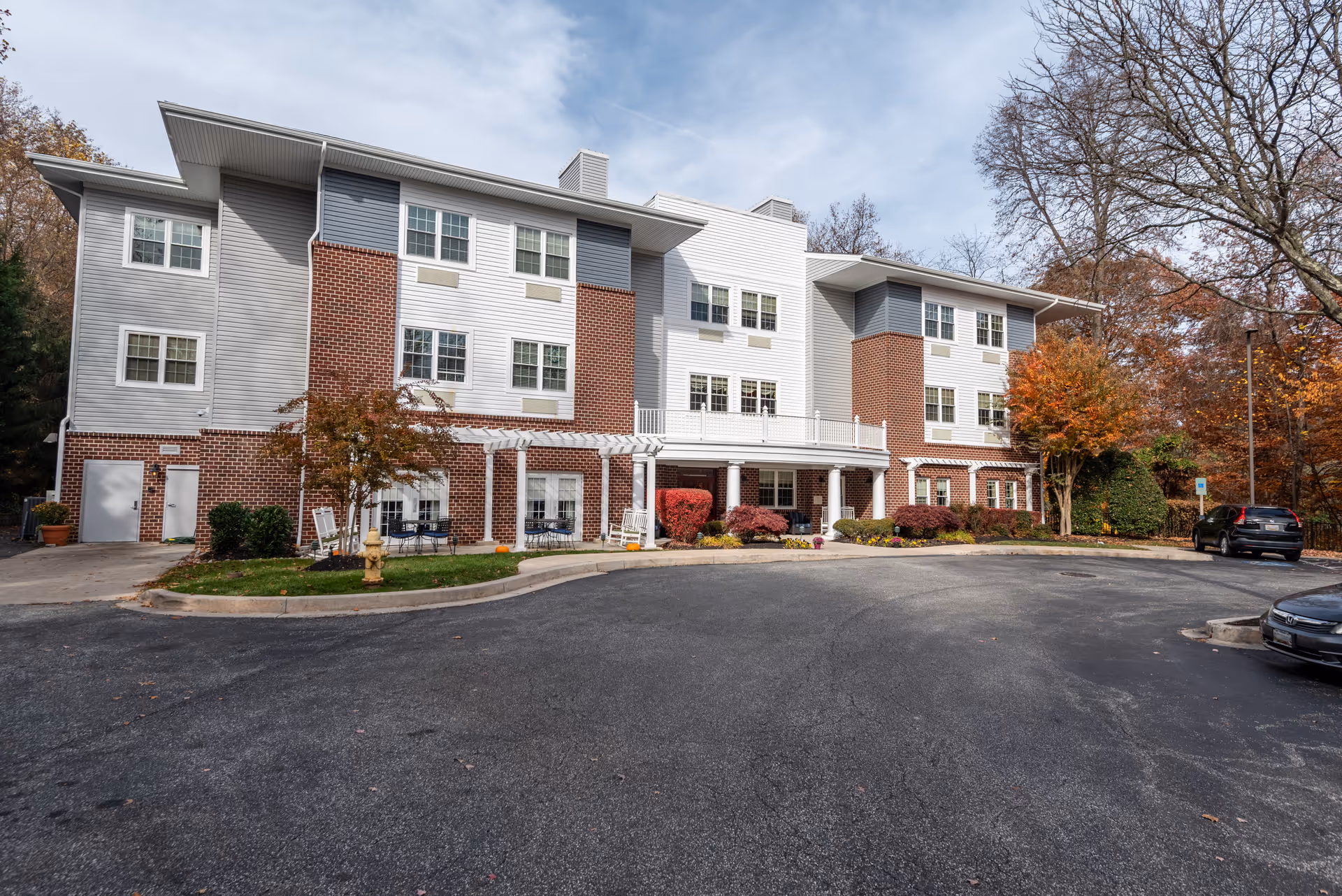 Three-story senior living building with white siding and red brick facade, a covered entrance, landscaped shrubs and parked cars in front under a partly cloudy sky.