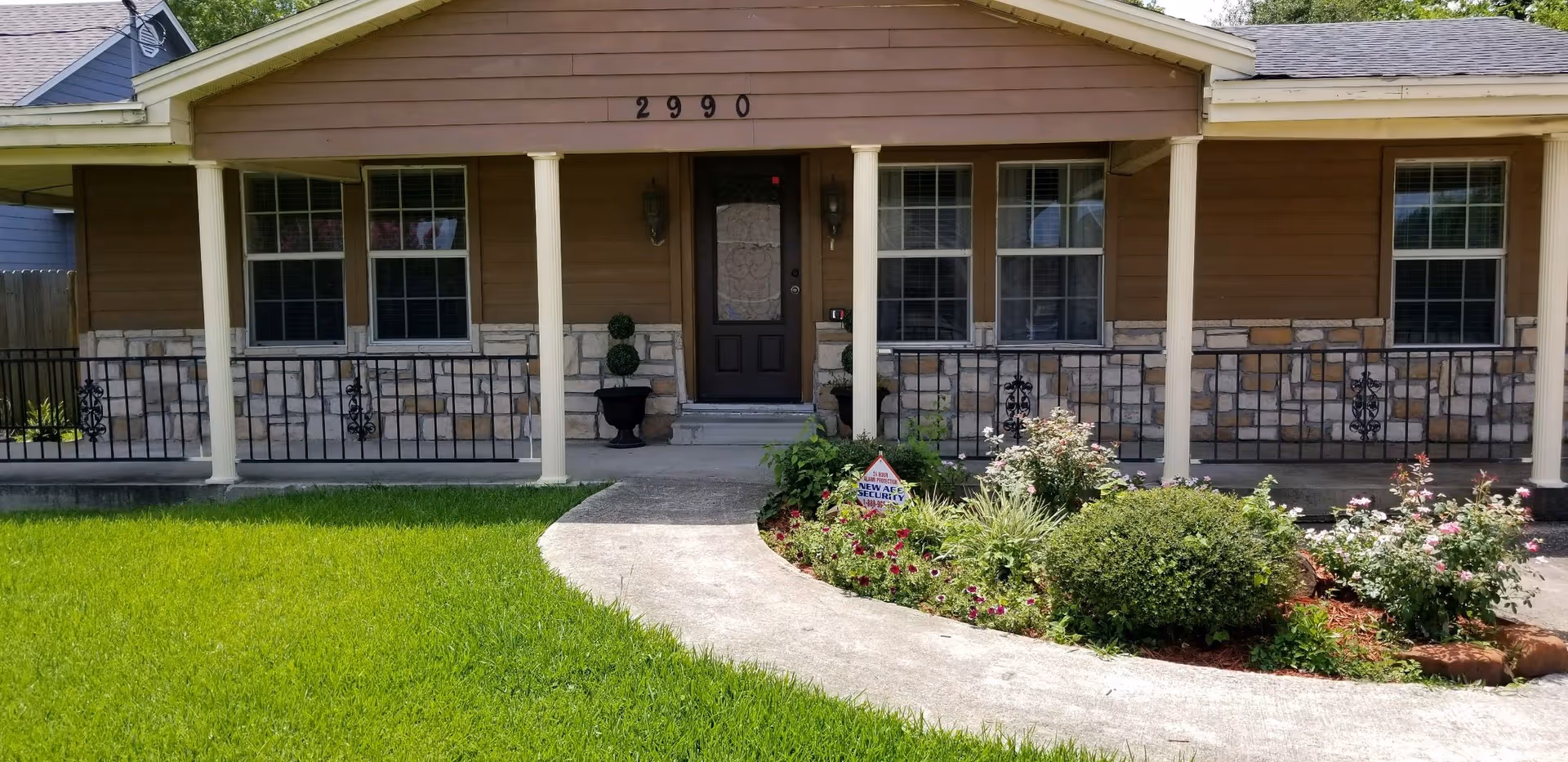 Front exterior view of a single-story building with brown siding and stone accents, featuring a covered porch with white columns, a dark front door, and several windows. The address number 2990 is displayed above the door. There is a curved concrete walkway leading through a well-maintained lawn and garden with various plants and flowers.