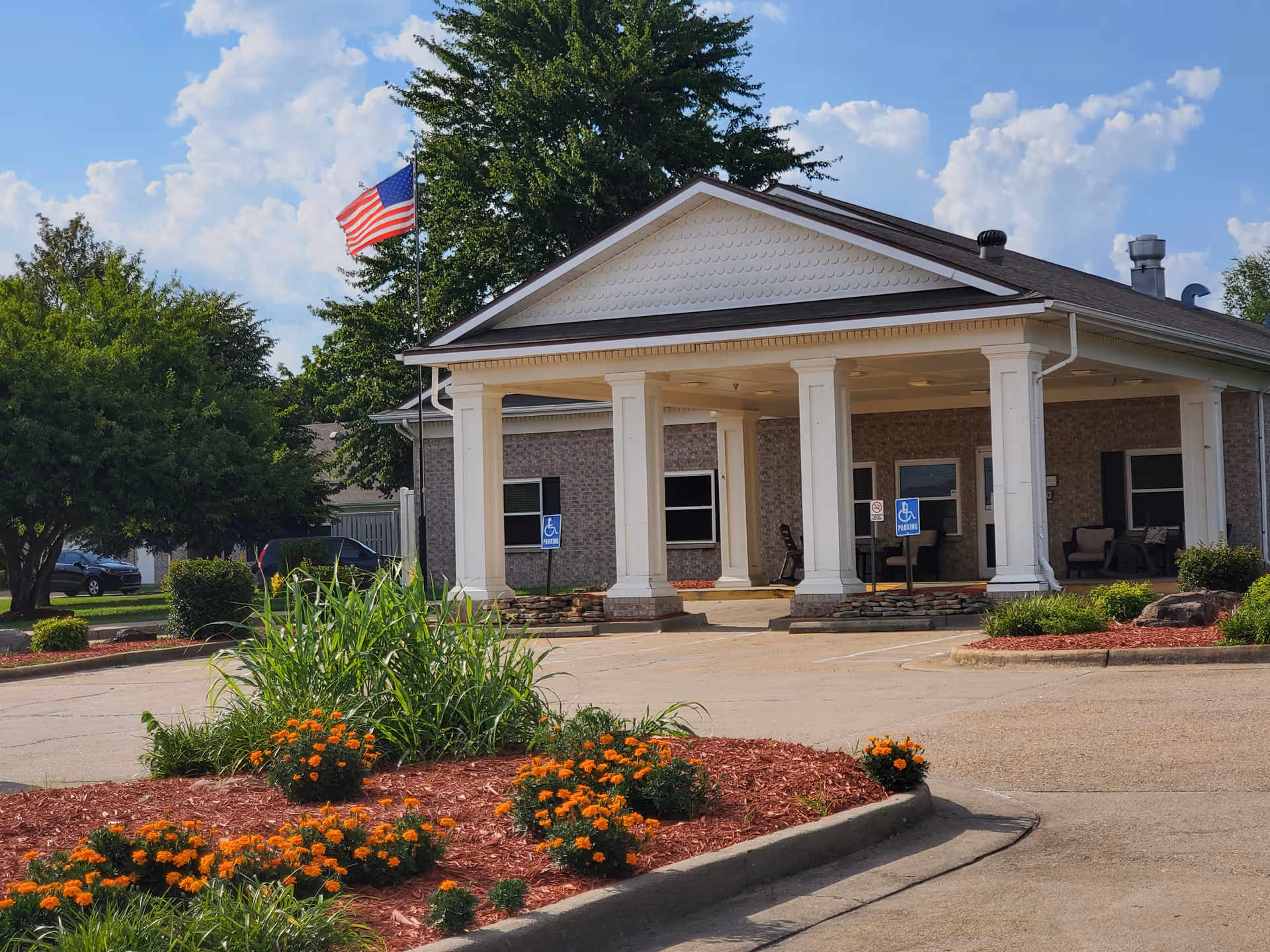 Front entrance of Autumn Hill Assisted Living with columns, flag, parking, and landscaped beds.