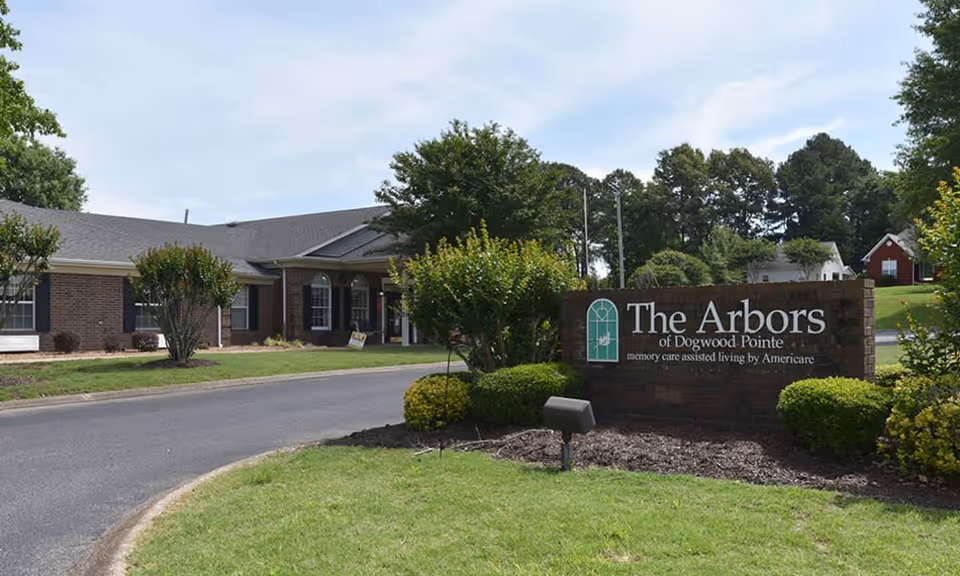Exterior view of The Arbors of Dogwood Pointe senior living facility with a brick building, a driveway, green lawns, bushes, and trees under a partly cloudy sky.
