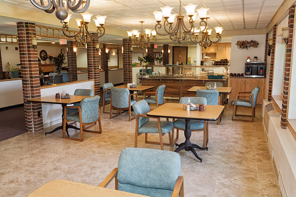 Dining room with multiple wooden tables and teal upholstered chairs, chandeliers, and a self-serve food counter in a senior living facility.