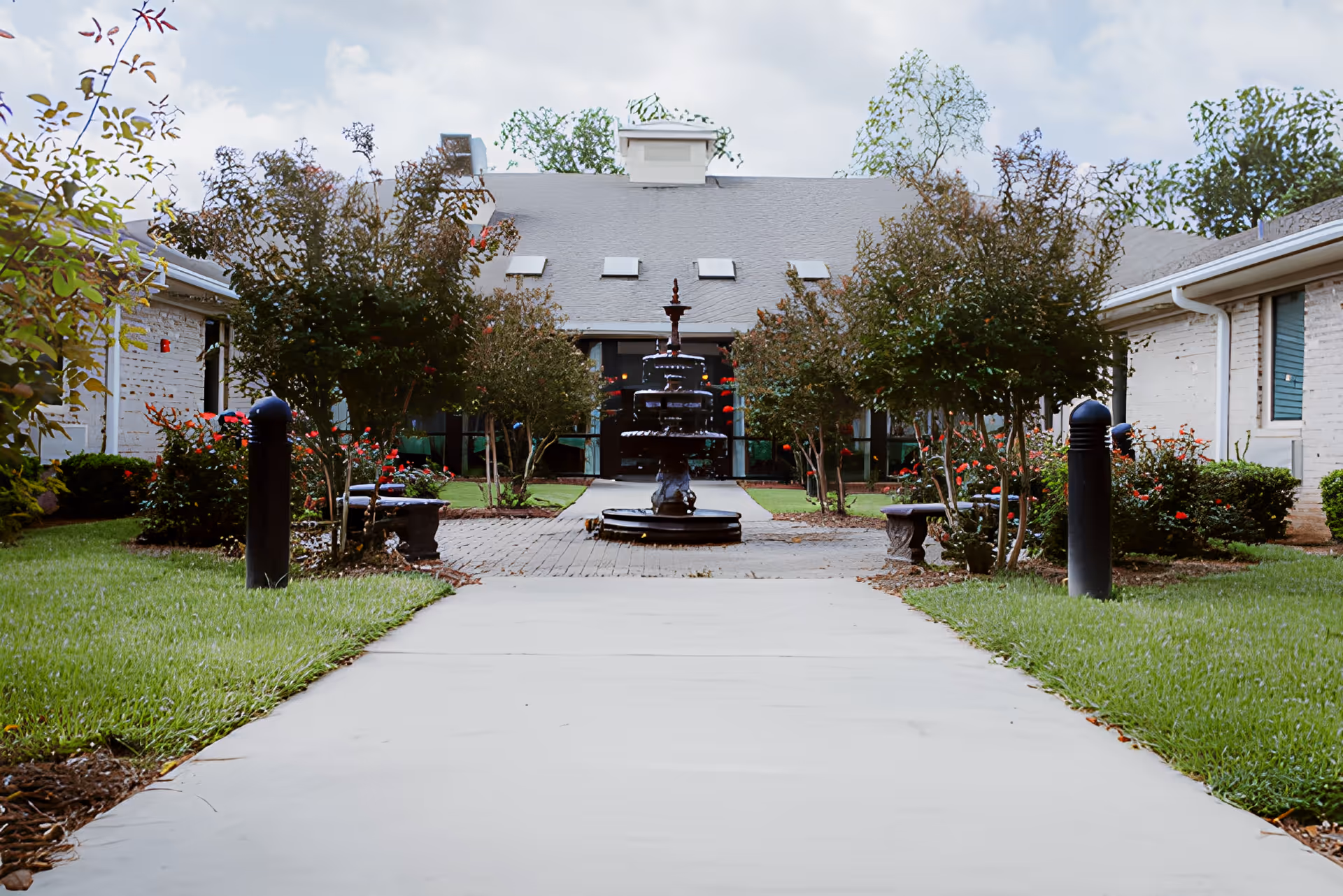 Concrete walkway leading through a landscaped courtyard to a building entrance with a central fountain and surrounding trees.