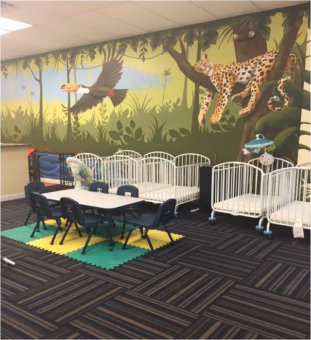 Bright indoor play area with white cribs along a jungle mural and small table with chairs.