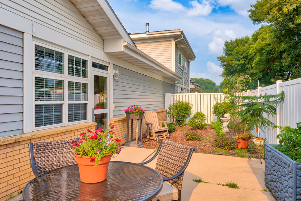 Outdoor patio area at Autumn Grace II featuring a round glass table with a potted plant of red flowers, two wicker chairs, a wooden Adirondack chair, and various potted plants and shrubs along a white fence under a partly cloudy sky.
