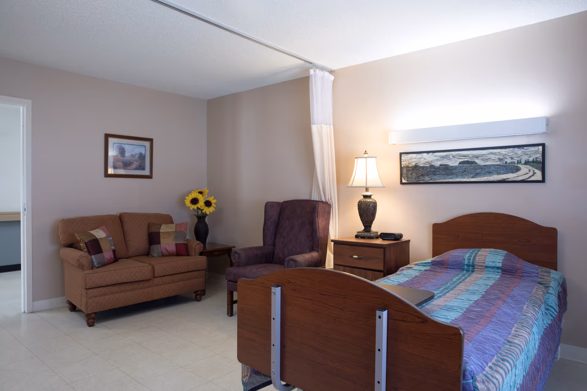 A senior living facility room with a single bed covered in a colorful striped blanket, a wooden nightstand with a lamp and a clock, a purple armchair, a brown loveseat with decorative pillows, a small side table with a vase of sunflowers, and framed artwork on the walls.