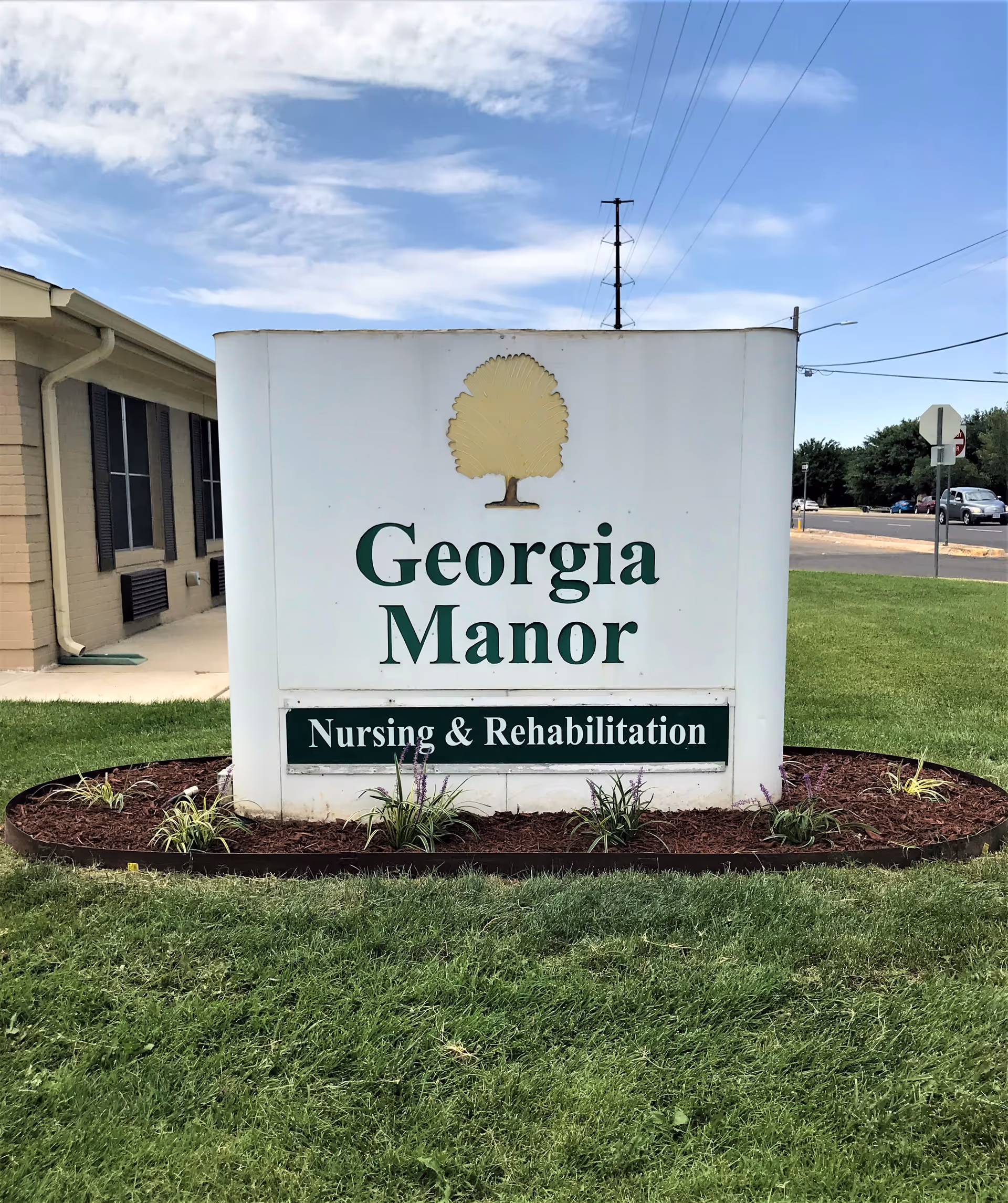 Outdoor sign for Georgia Manor Nursing & Rehabilitation facility with a tree logo, situated on a grassy area with a building and street visible in the background under a partly cloudy sky.
