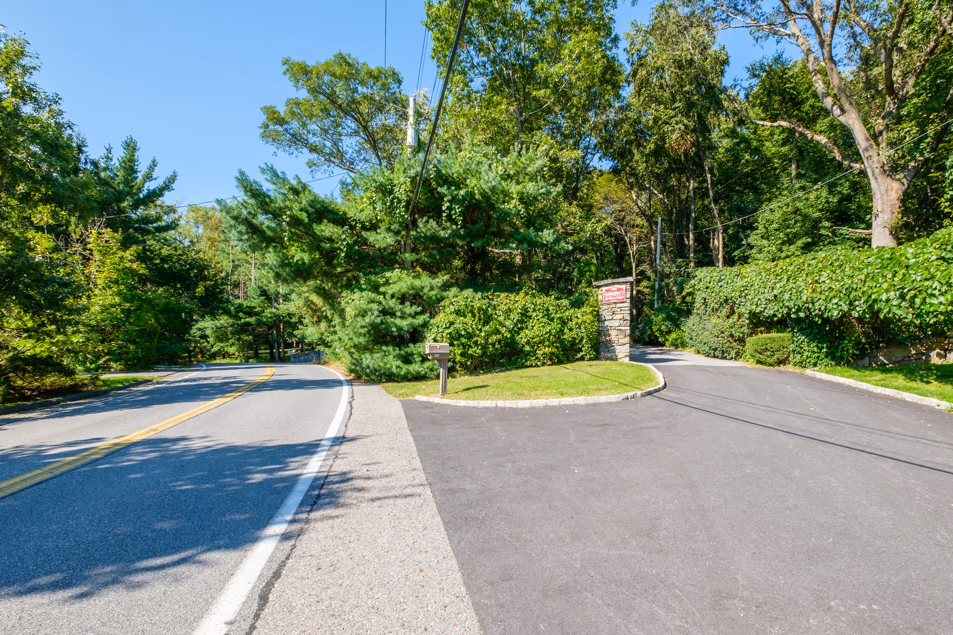 A paved road curves to the left with a driveway branching off to the right leading into a wooded area. There is a stone pillar with a red sign that reads 'Gallagher Home' at the entrance of the driveway. The scene is surrounded by green trees and bushes under a clear blue sky.