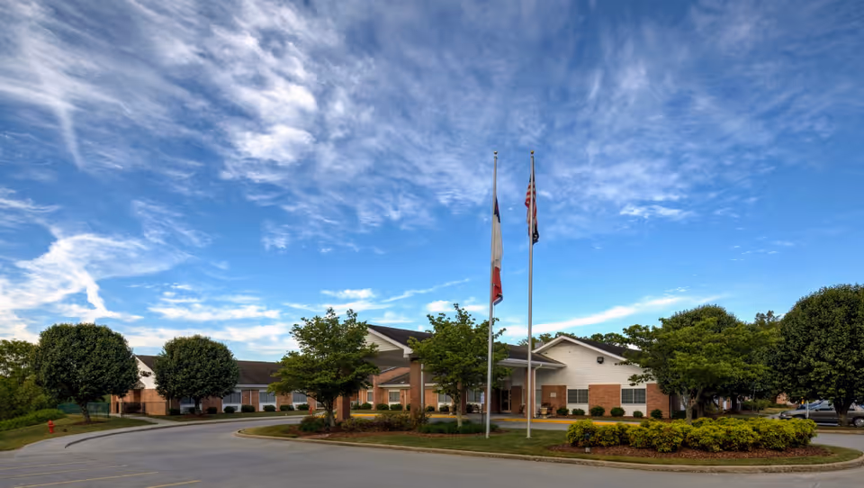 Brick senior living facility with curved driveway, flagpoles, and trees under a blue sky.