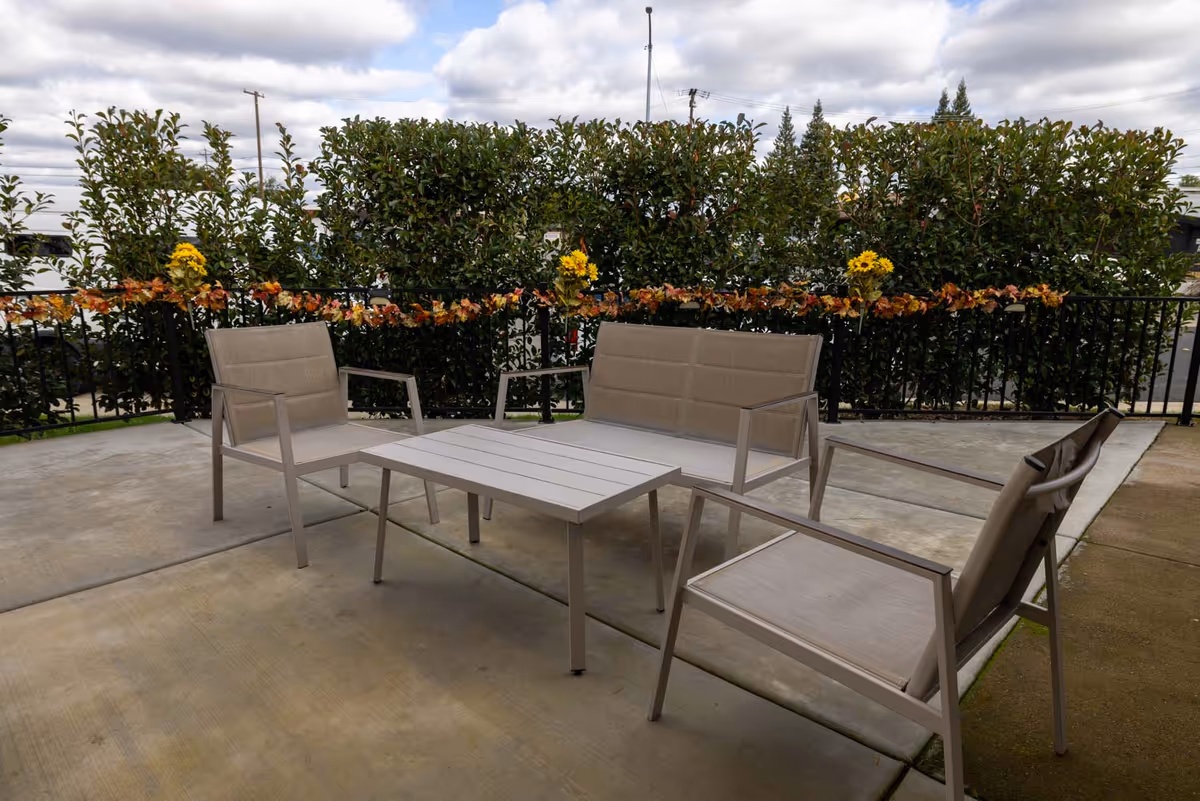 Outdoor patio with beige chairs and table surrounded by hedges and garland.