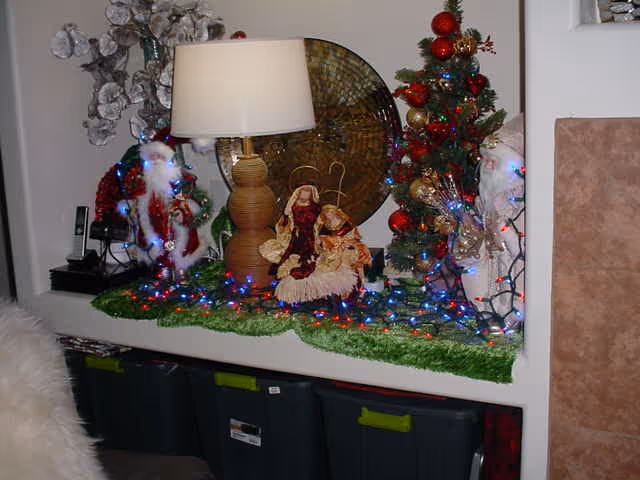 A festive holiday display on a shelf featuring a table lamp, a nativity scene with Mary and baby Jesus, two Santa Claus figurines, a small decorated Christmas tree, colorful string lights, and a decorative round wall piece in the background.