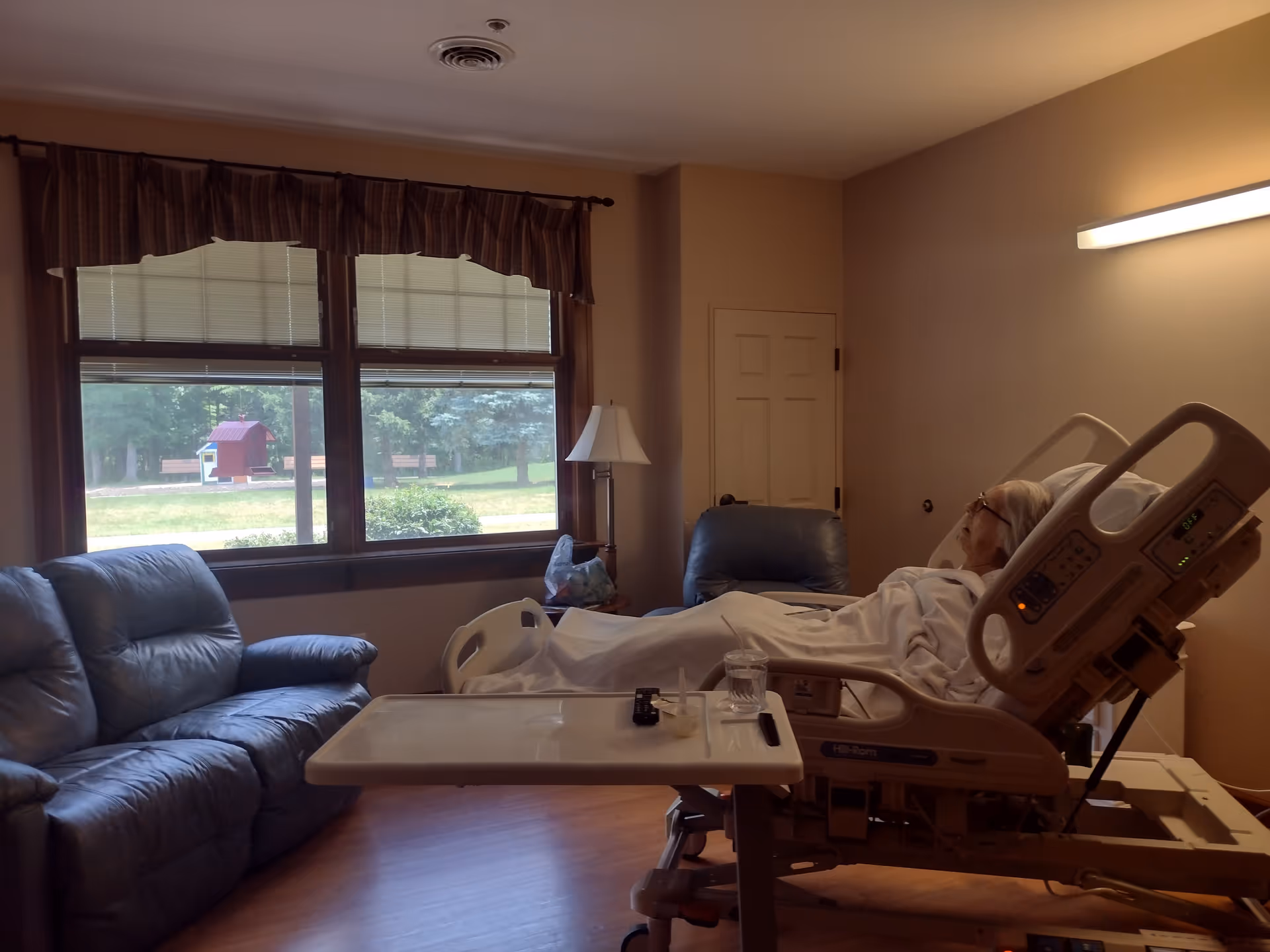 Senior patient rests in a hospice room with a hospital bed, sofa, and large window.