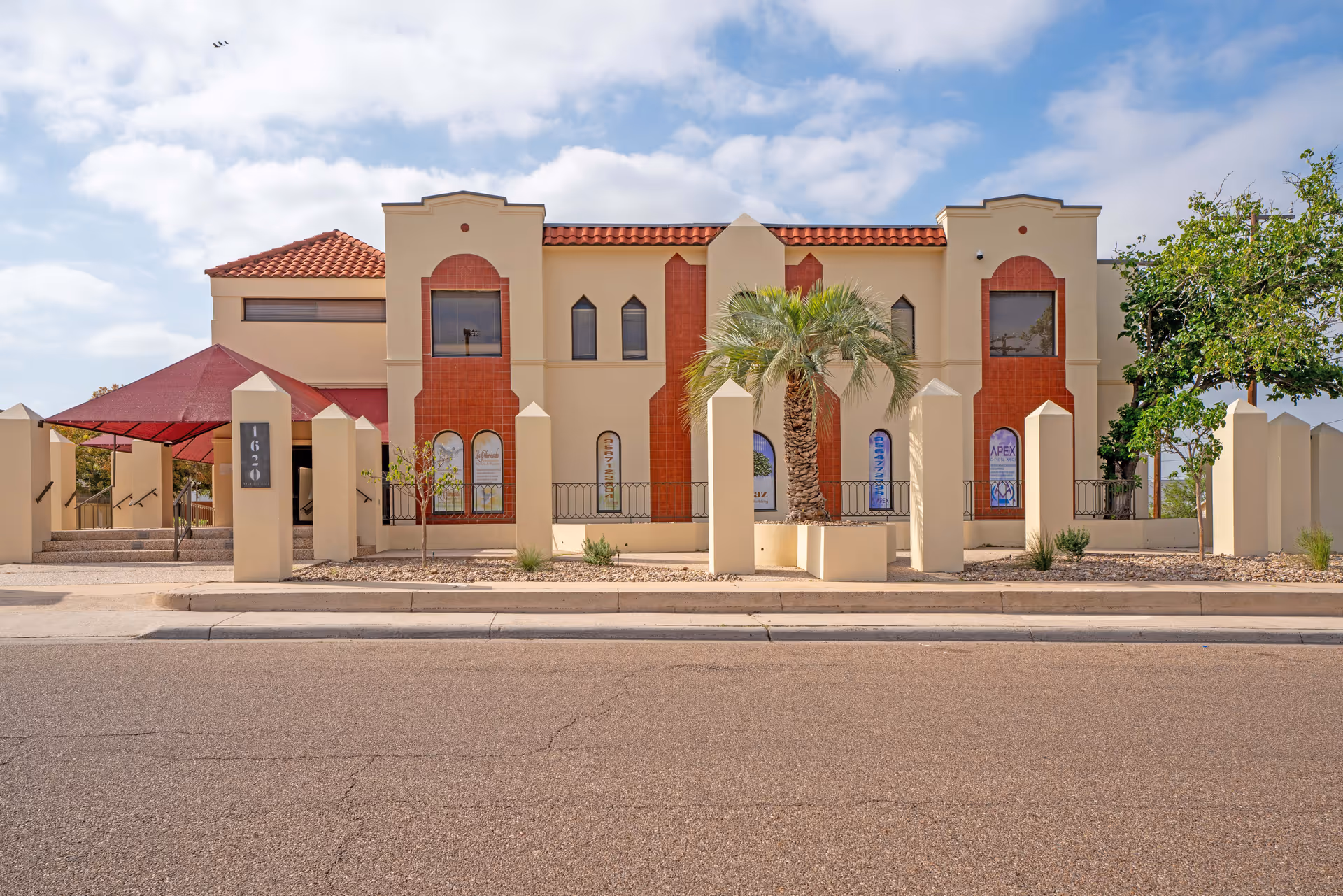 Beige building with red-tiled roof, palm trees, and a gated front along a street.