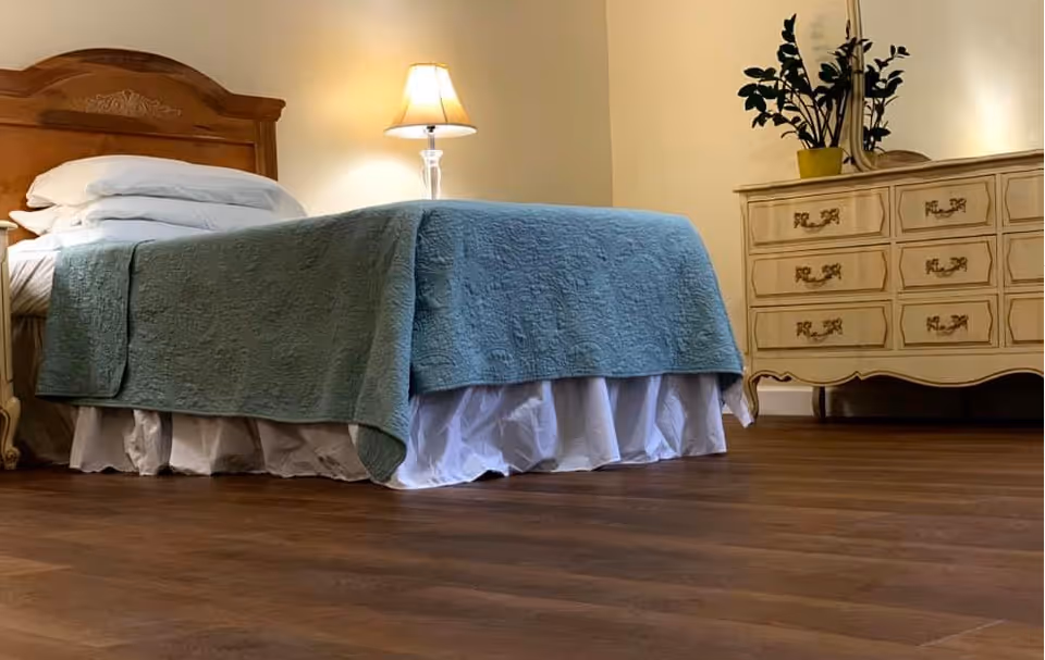 Cozy bedroom featuring a wooden headboard bed, blue quilt, dresser, lamp, and wooden floor.
