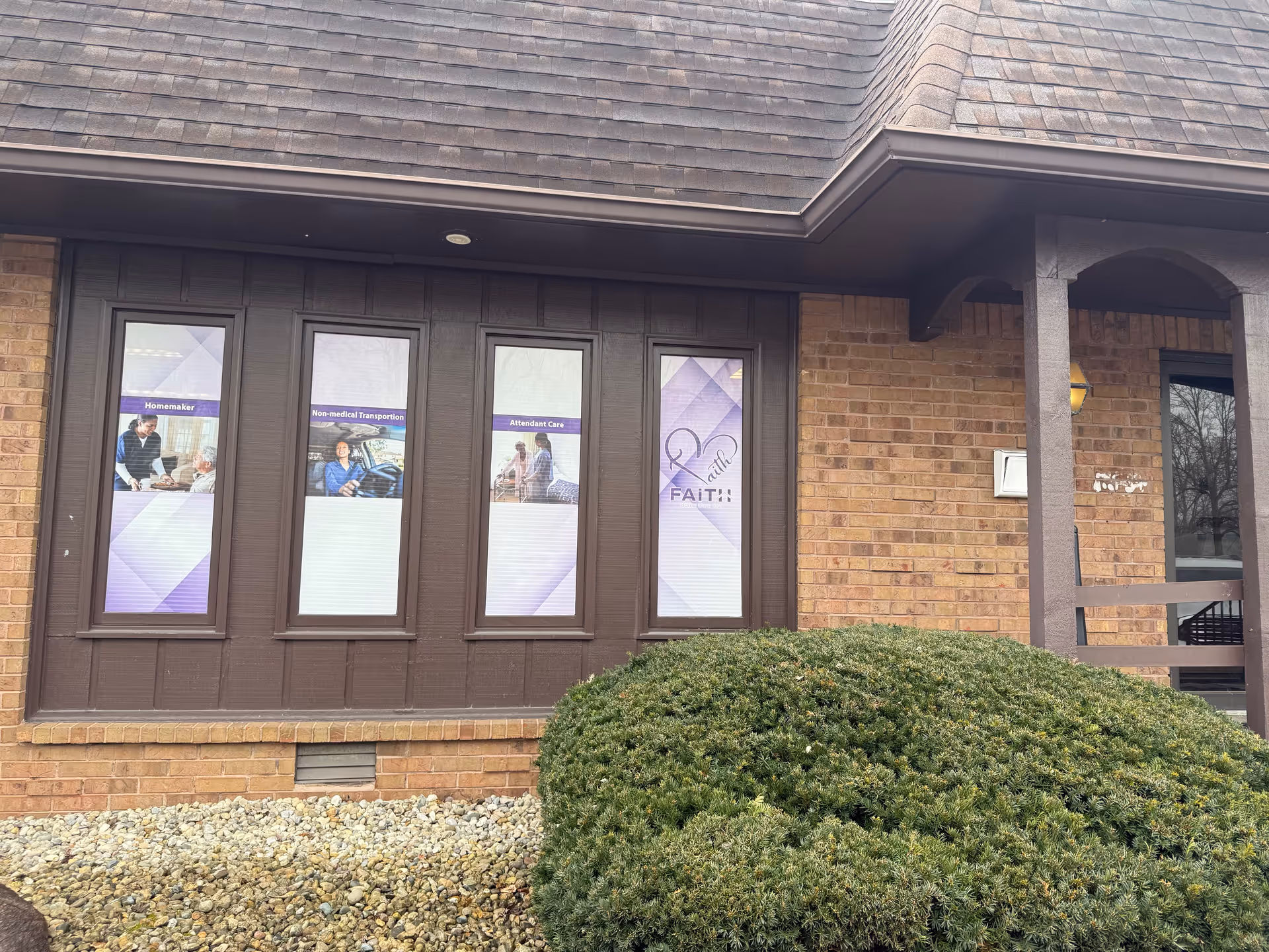 Front exterior of a brick senior care facility with purple window banners and shrubs.