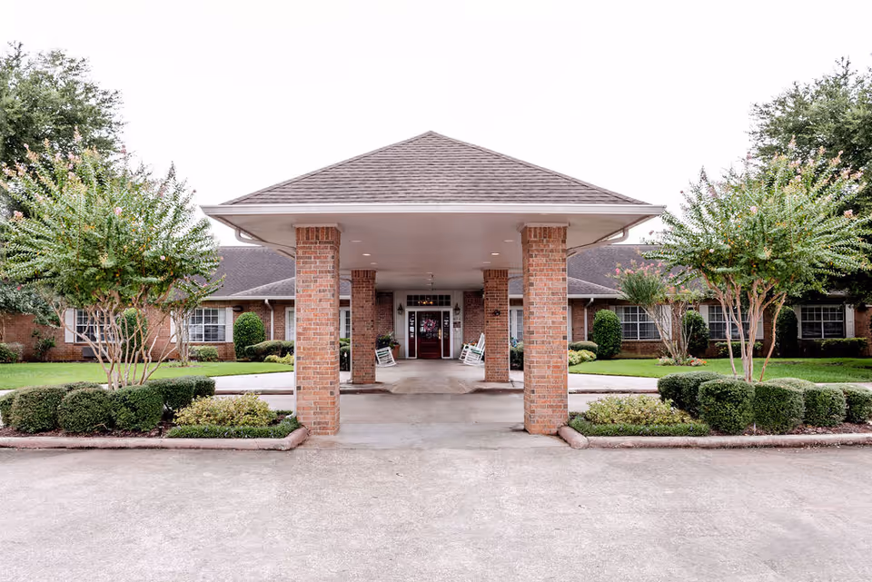 Front entrance of The Reserve at First Colony Assisted Living and Memory Care facility featuring a covered driveway supported by brick columns, with manicured bushes and trees on either side and a brick building in the background.