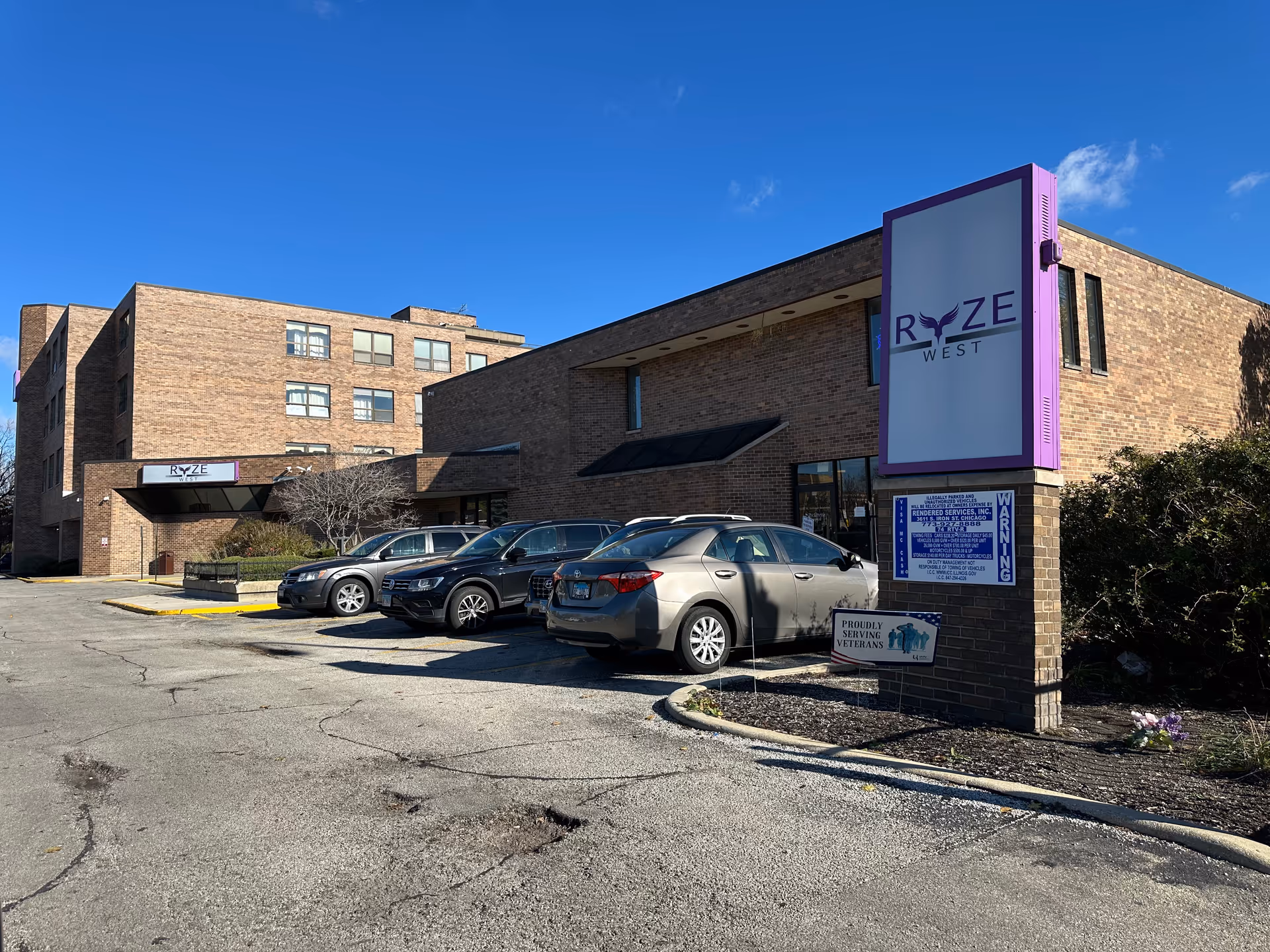 Front exterior of the RYZE West senior living building with its sign, parked cars, and a clear blue sky.