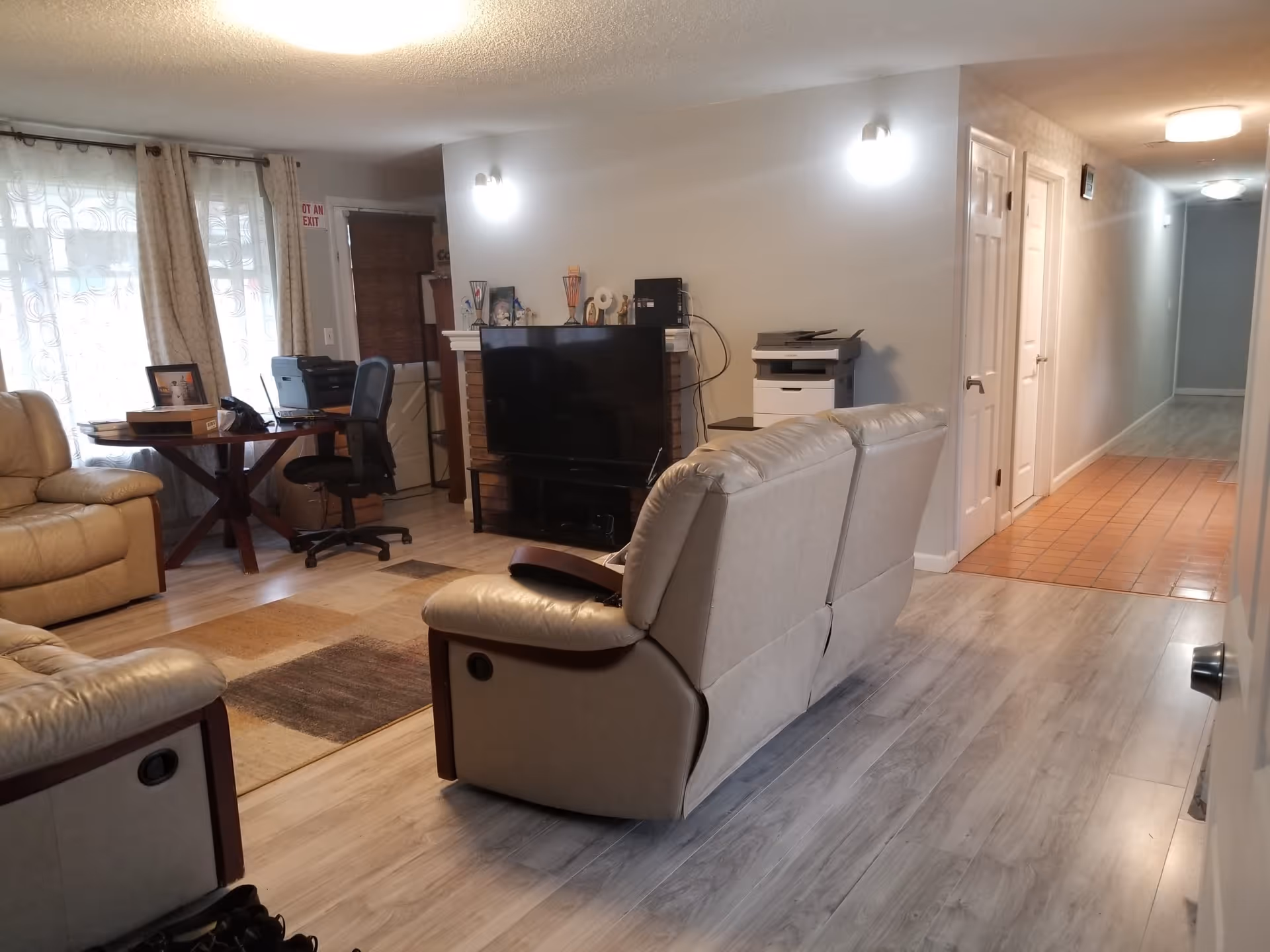Interior view of a living room area in Trinity Adult Care Home featuring beige leather recliners, a flat-screen TV on a stand, a small office desk with a chair and printer near a window with curtains, and a hallway with tiled flooring leading to other rooms.