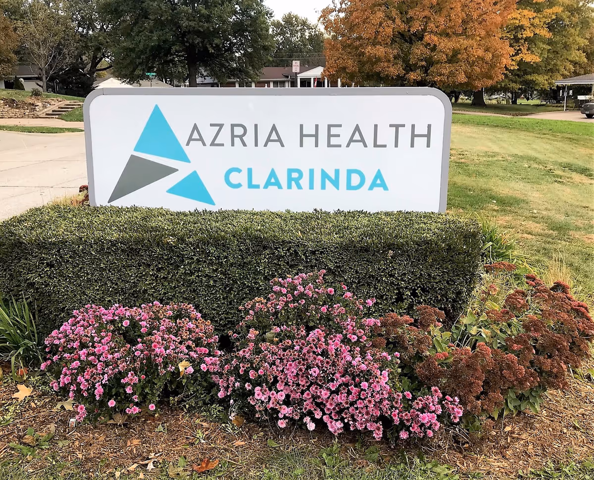 Large outdoor sign reads Azria Health Clarinda with triangular logo, framed by shrubs and flowers.
