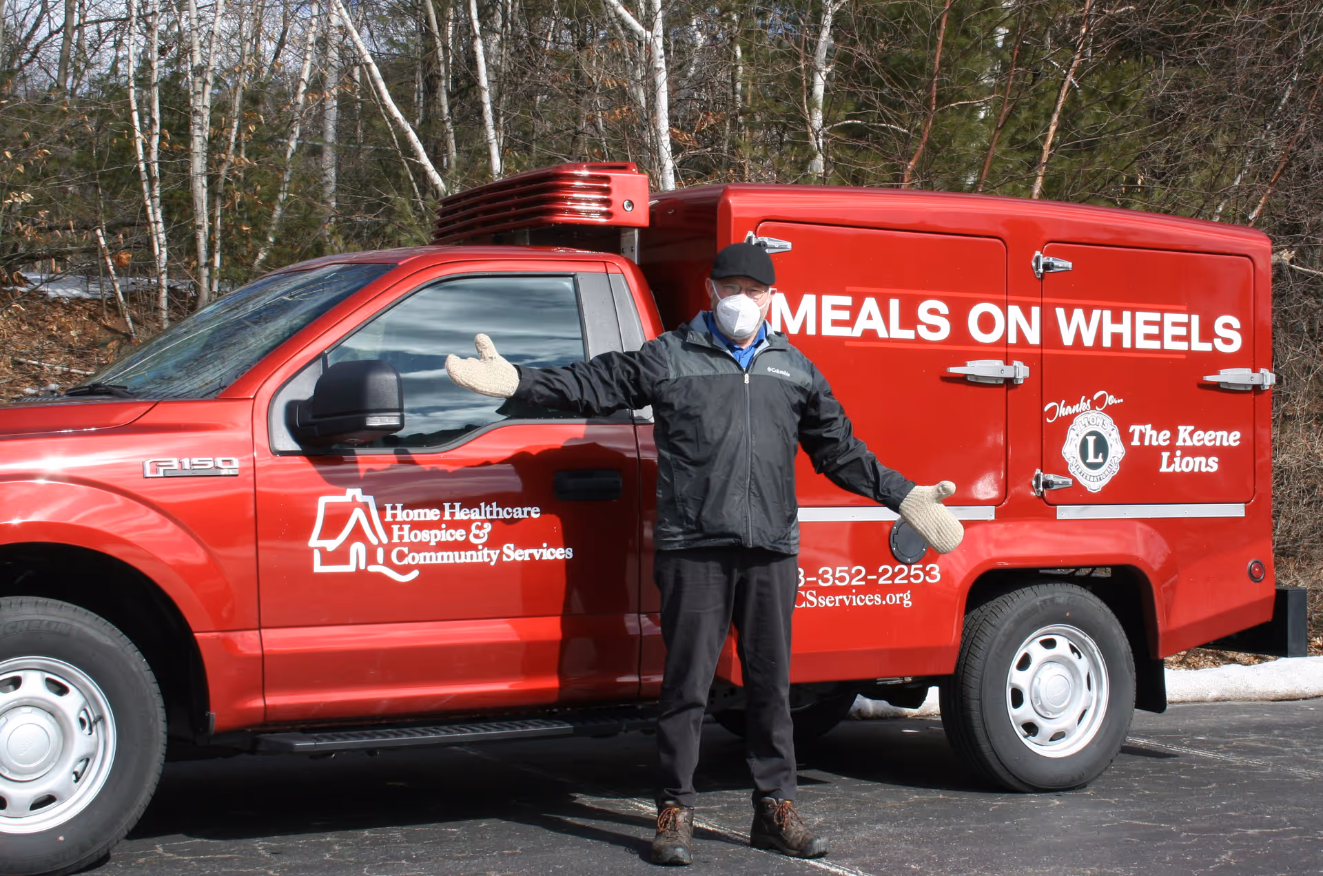 A masked worker stands beside a red Meals on Wheels van in a parking lot.