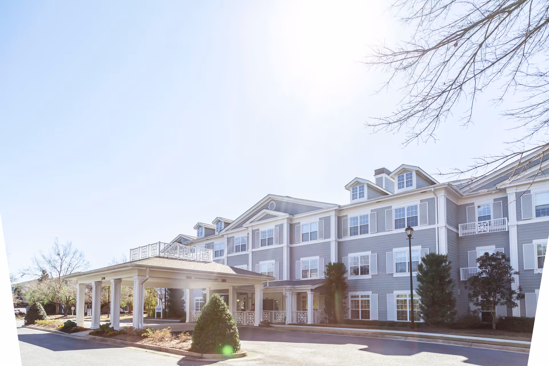 Exterior view of a large, three-story senior living facility building with gray siding and white trim. The building features multiple windows, dormer windows on the roof, and a covered entrance supported by white columns. There are trees and shrubs around the building, and the sky is clear and sunny.