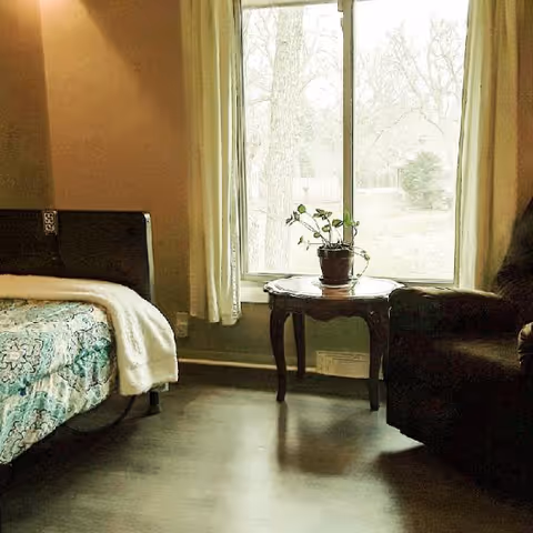 A cozy bedroom with a bed on the left covered with a patterned blue and white blanket and a white throw. Next to the bed is a small wooden table with a potted plant on it, positioned in front of a large window with light-colored curtains. On the right side, there is a dark upholstered armchair. The room has wooden flooring and soft lighting.