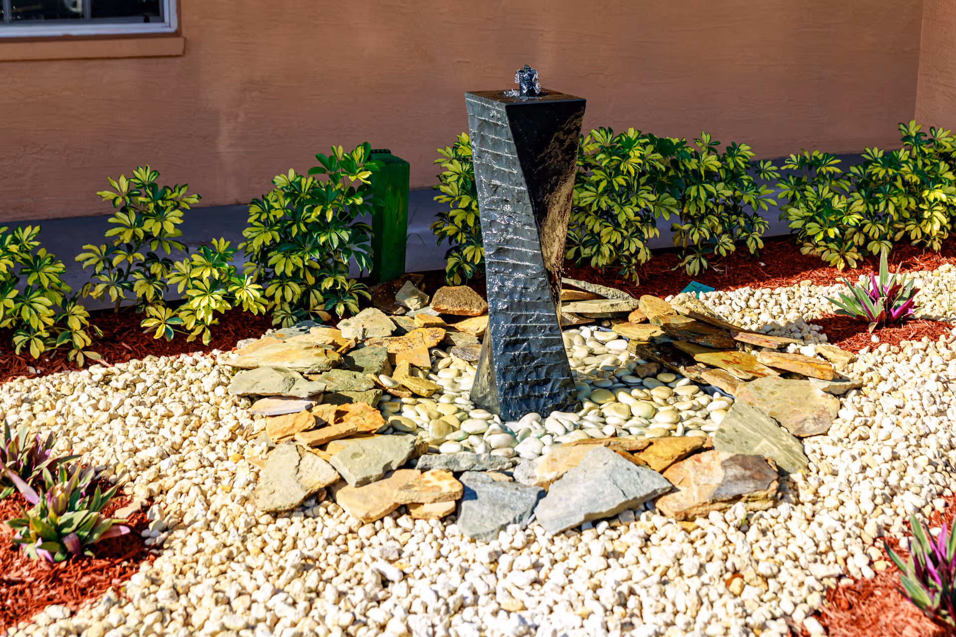 Stone fountain centerpiece surrounded by rocks and shrubs in a courtyard beside a peach wall.