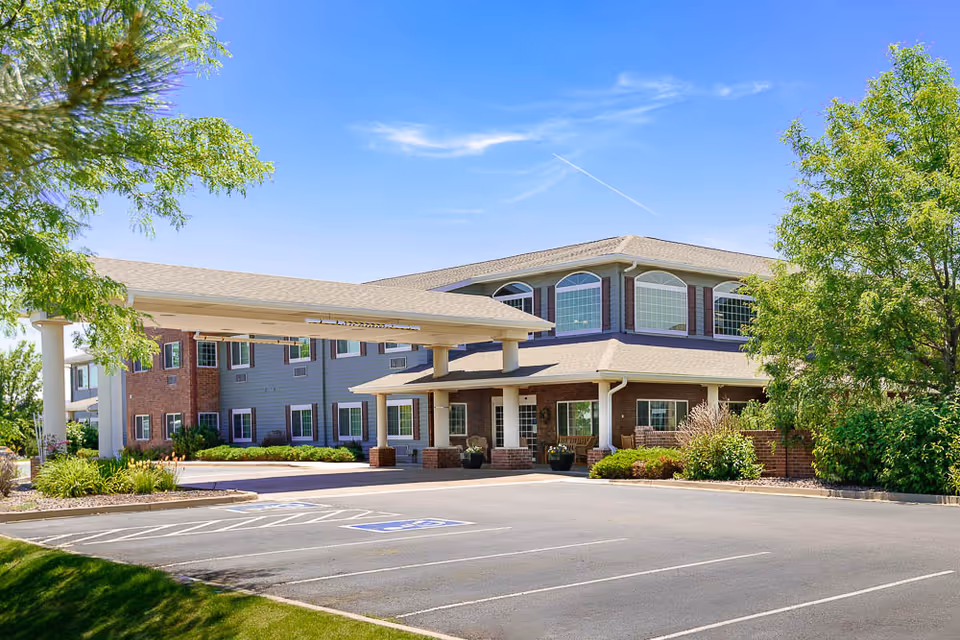 Front exterior of a two-story senior living facility with a covered entrance, parking lot, and landscaped trees.