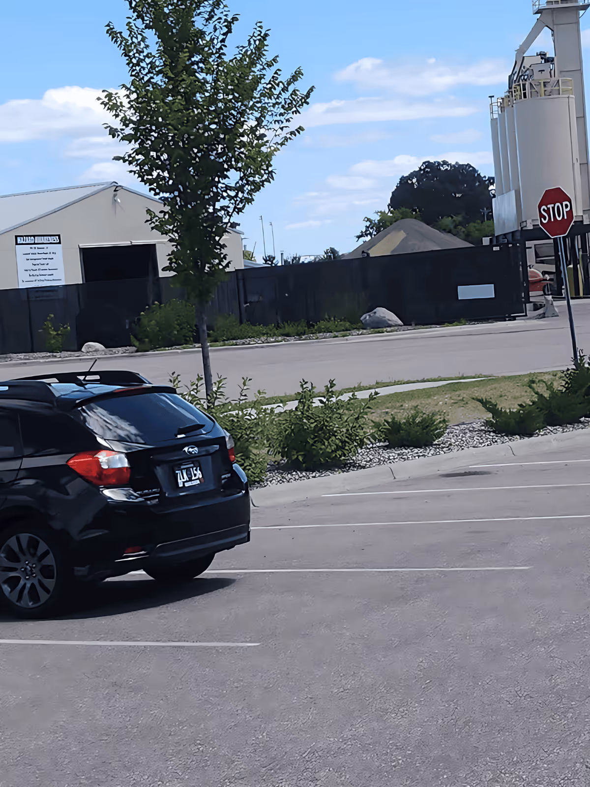 Parking lot scene with a black SUV, trees, and industrial buildings in the background.