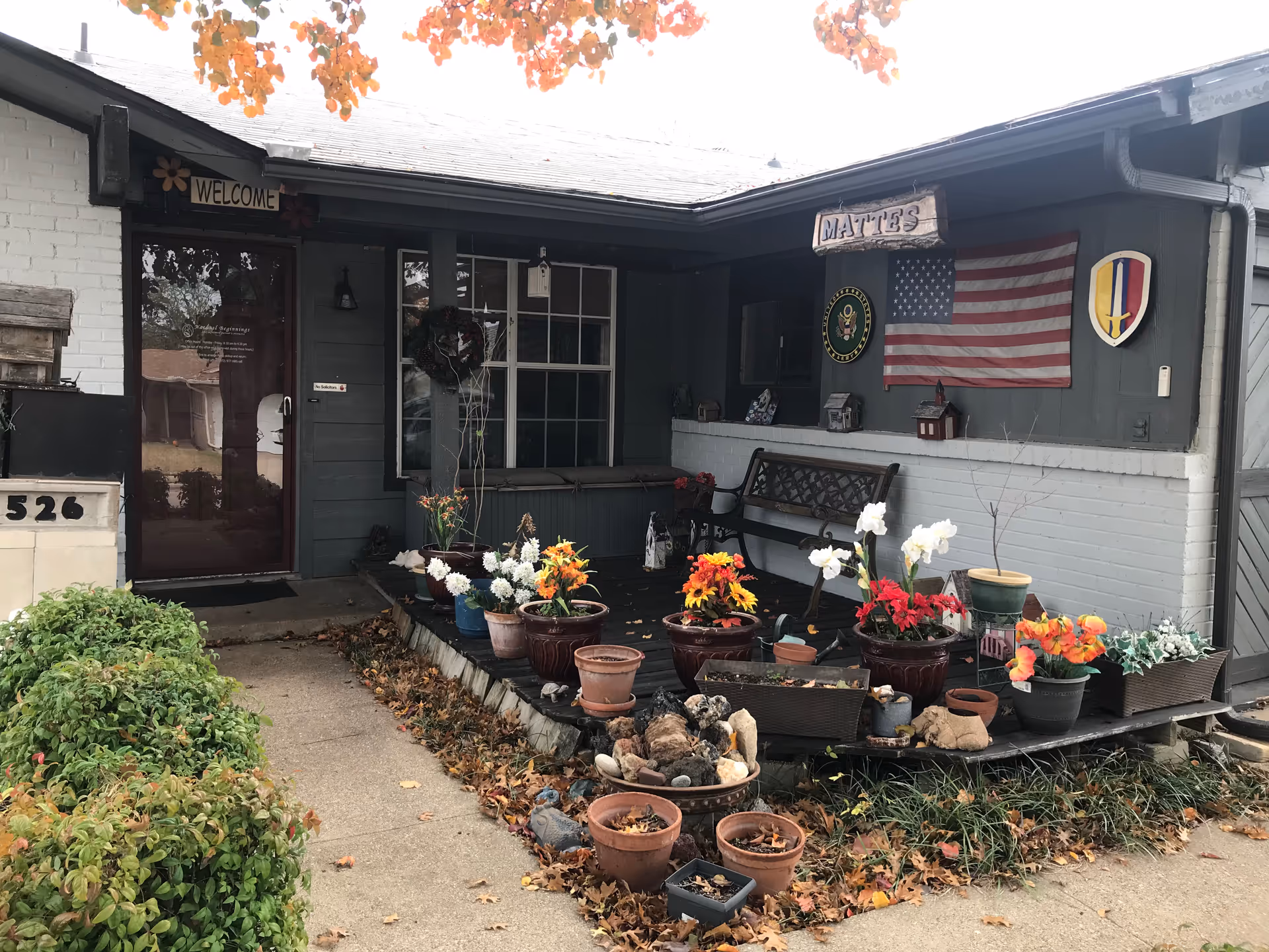 Front porch of a senior living facility with potted flowers, bench, and a flag display.