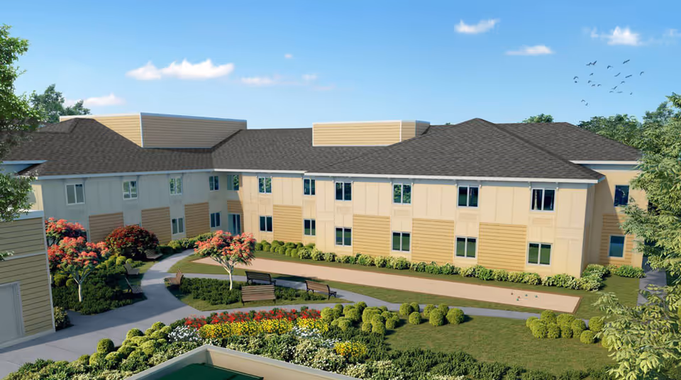 Exterior view of a senior living facility building with beige and light yellow siding, dark shingled roof, and multiple windows. The landscaped courtyard features walking paths, benches, green bushes, and flowering trees under a blue sky with scattered clouds and birds flying.
