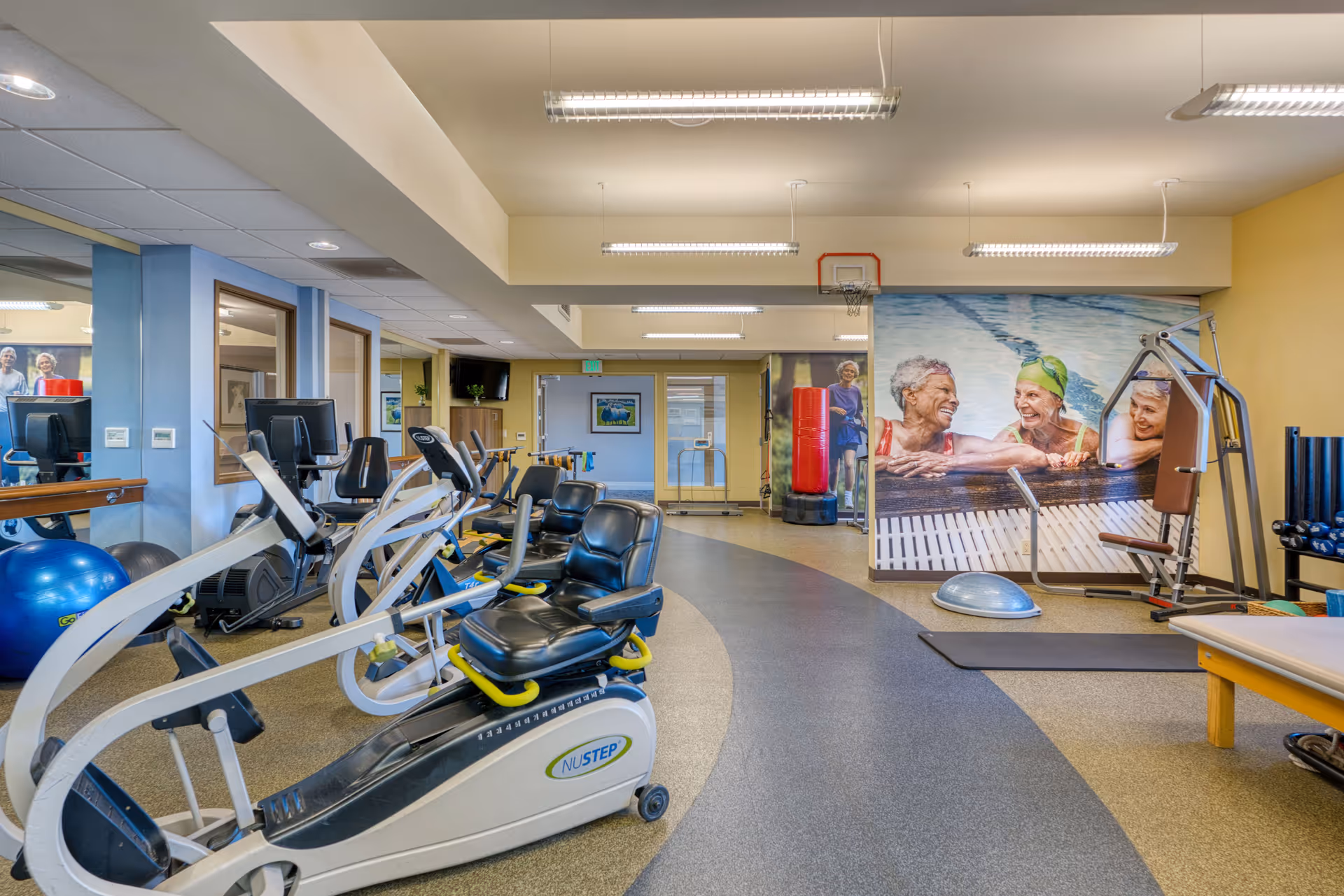 Senior fitness center with cardio machines, mats, and mural on yellow walls.