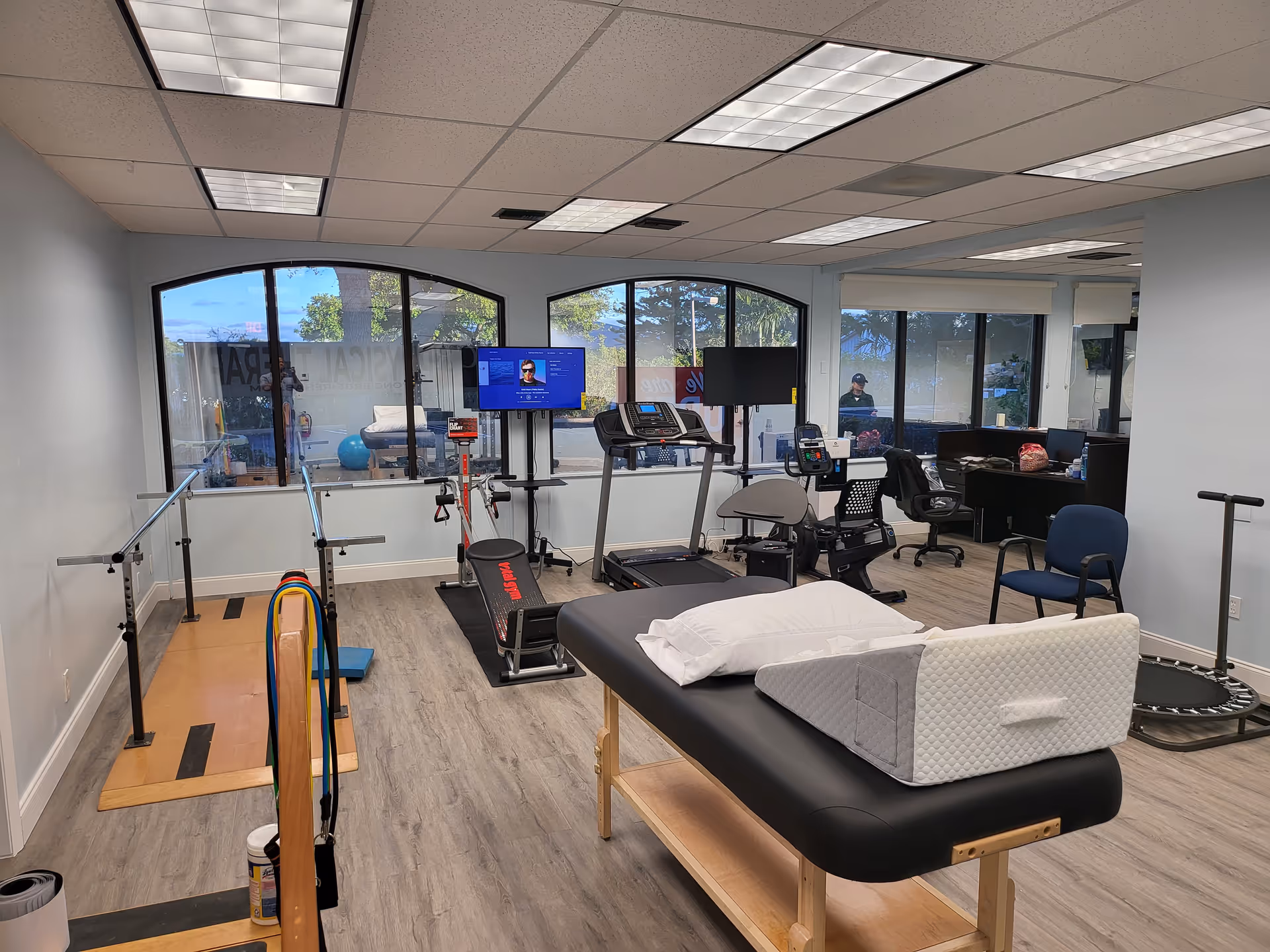 Therapy gym with treatment table, parallel bars, treadmills, and exercise equipment inside a rehab facility.