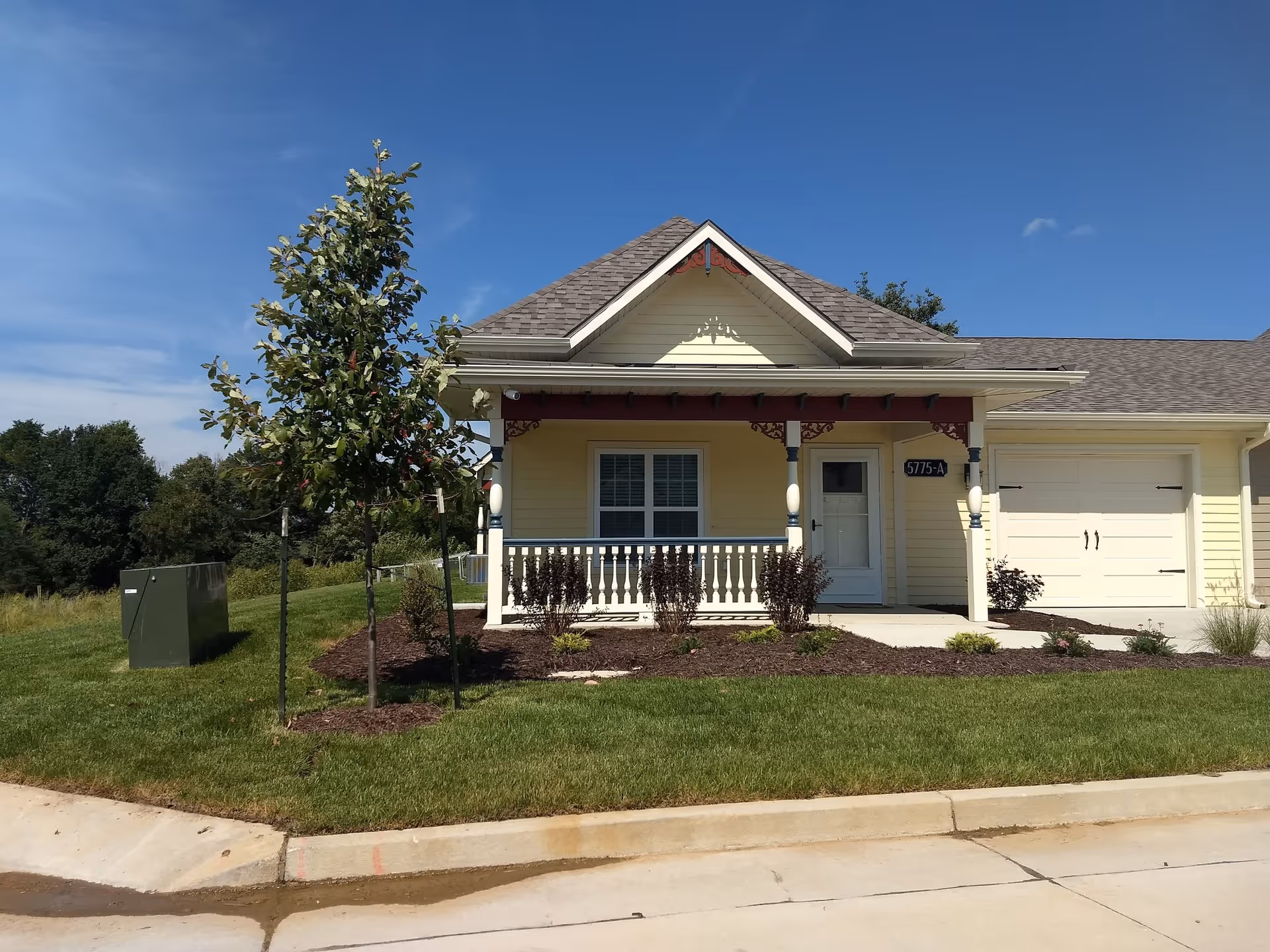 Front exterior view of a single-story yellow house with a small porch, white railing, and a garage. There is a young tree and some landscaping with bushes in front of the house under a clear blue sky.